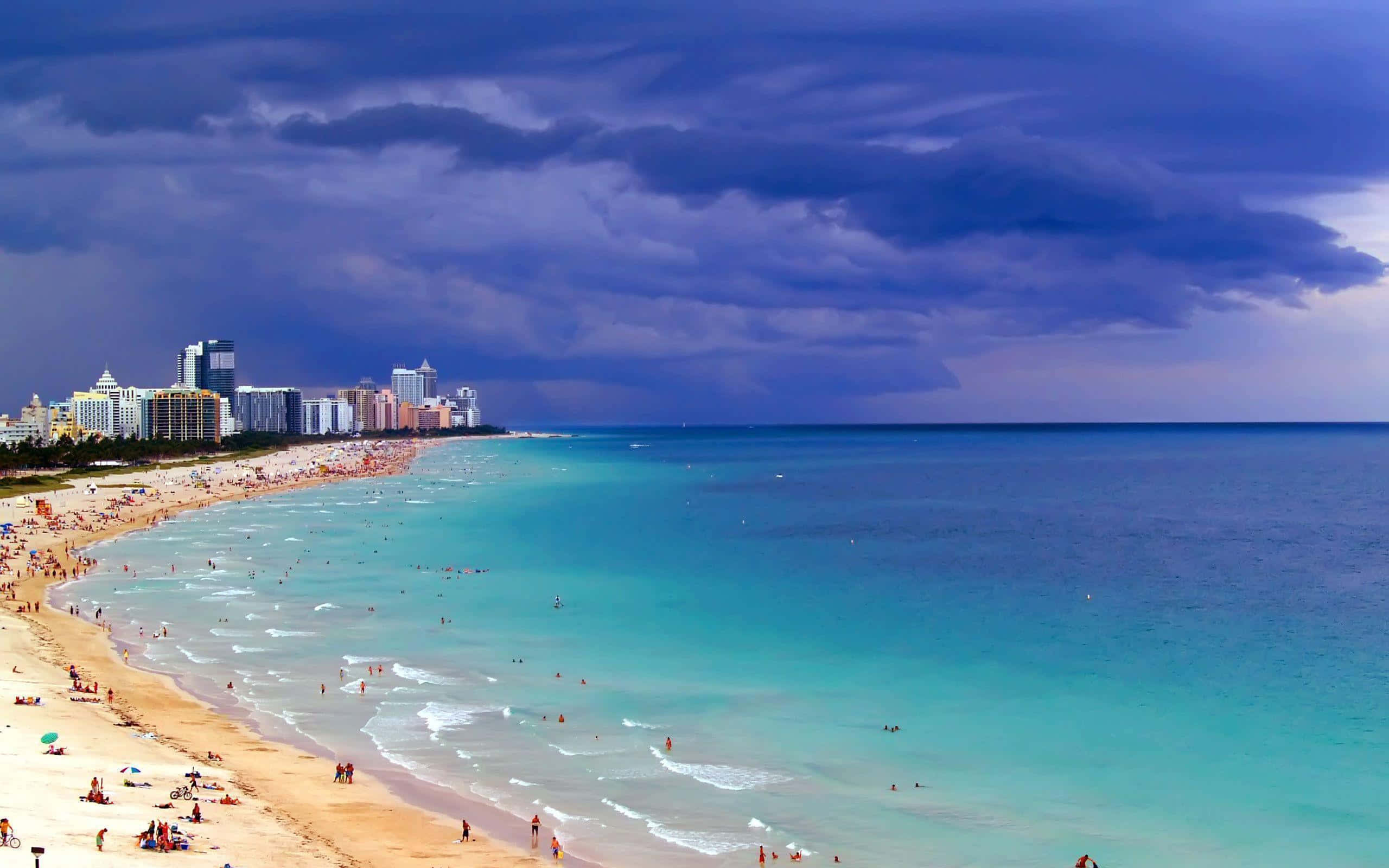 Miami 桌布 壁紙 — 充滿活力的海灘場景，有碧綠的海水和戲劇性的紫色暴風雲籠罩著擁擠的海岸線和城市天際線。