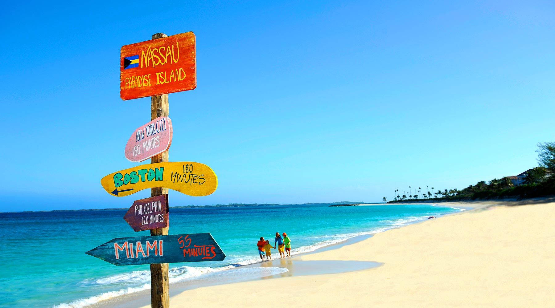 Nassau Bahamas 桌布 壁紙 — 充滿活力的海灘場景，色彩繽紛的方向標誌指向全球目的地，家庭在清澈藍天下沿著碧綠海水漫步。