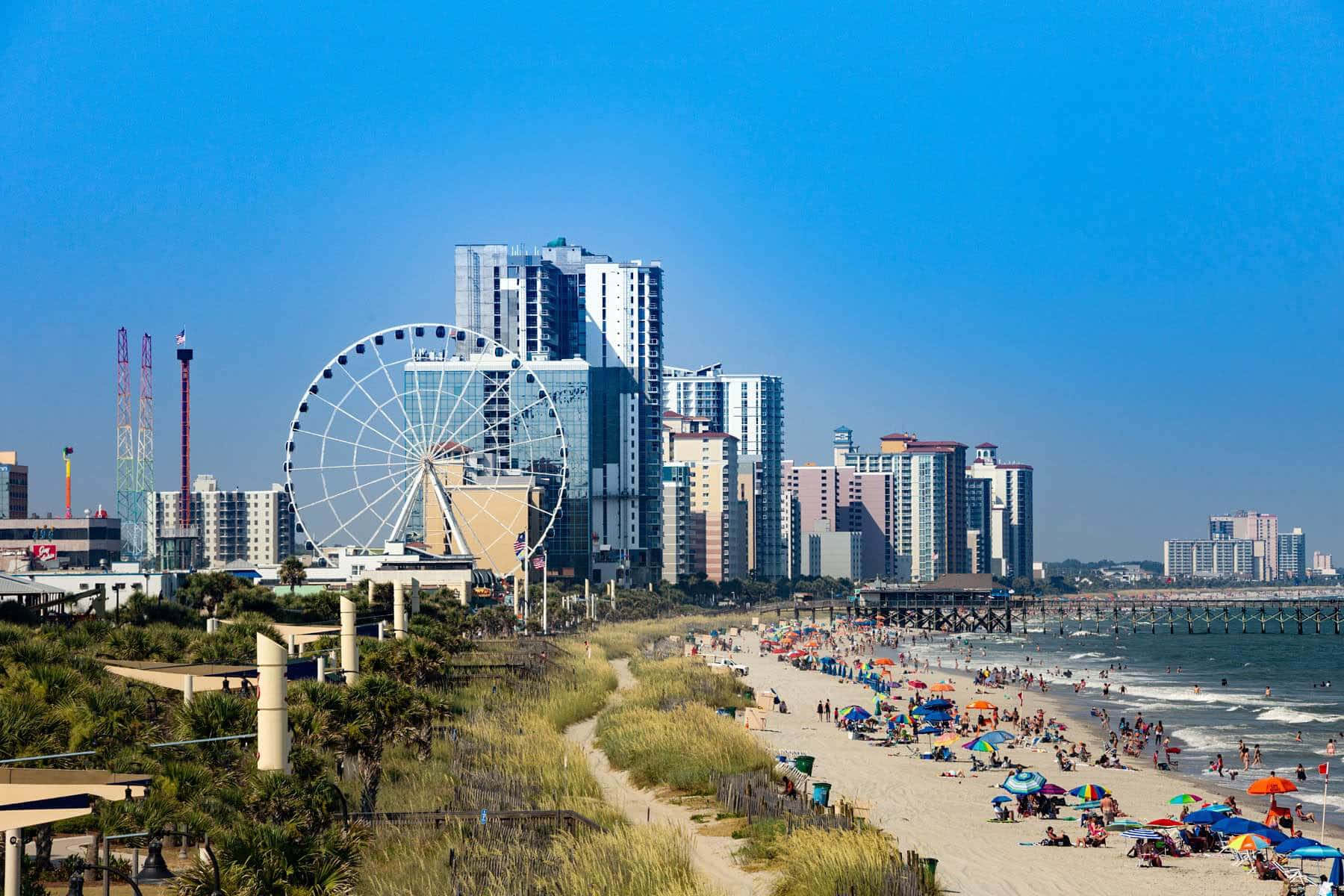 Myrtle Beach 桌布 壁紙 — 充滿活力的海灘場景，色彩繽紛的遮陽傘和人群，在晴朗的藍天下，旁邊是高聳的摩天輪和現代城市天際線。