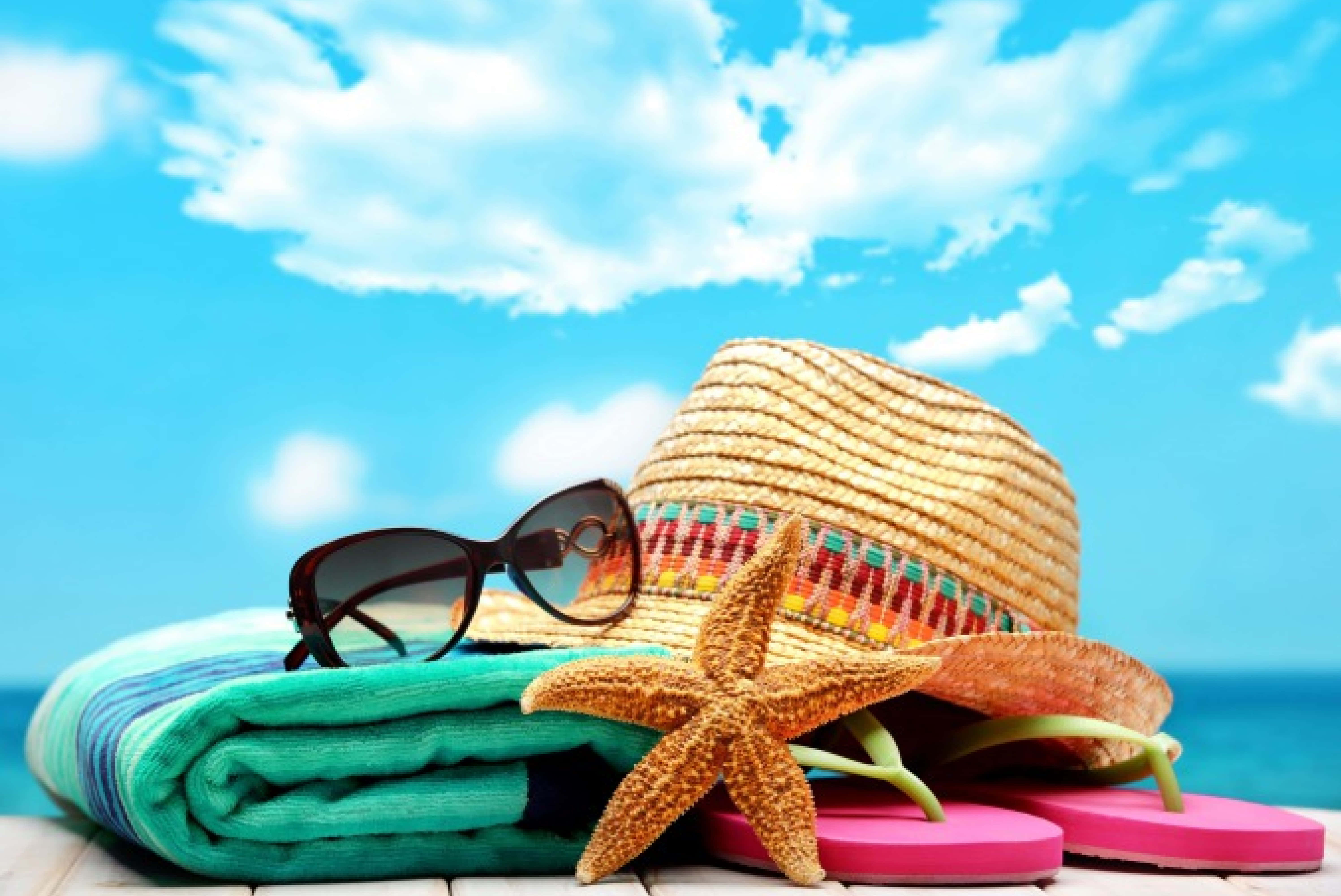 Beach Accessories 桌布 壁紙 — 充滿活力的海灘場景，草帽、太陽眼鏡、海星和粉紅色夾腳拖放在摺疊的毛巾上，上方是明亮的藍天。