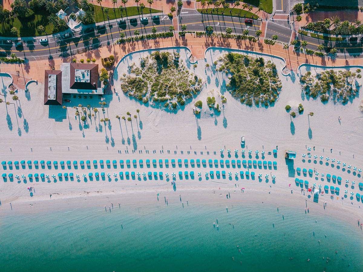 florida 桌布 壁紙 — 充滿活力的海灘鳥瞰圖，有碧綠的海水、一排排藍色遮陽傘和棕櫚樹在沙灘上投下長長的影子。