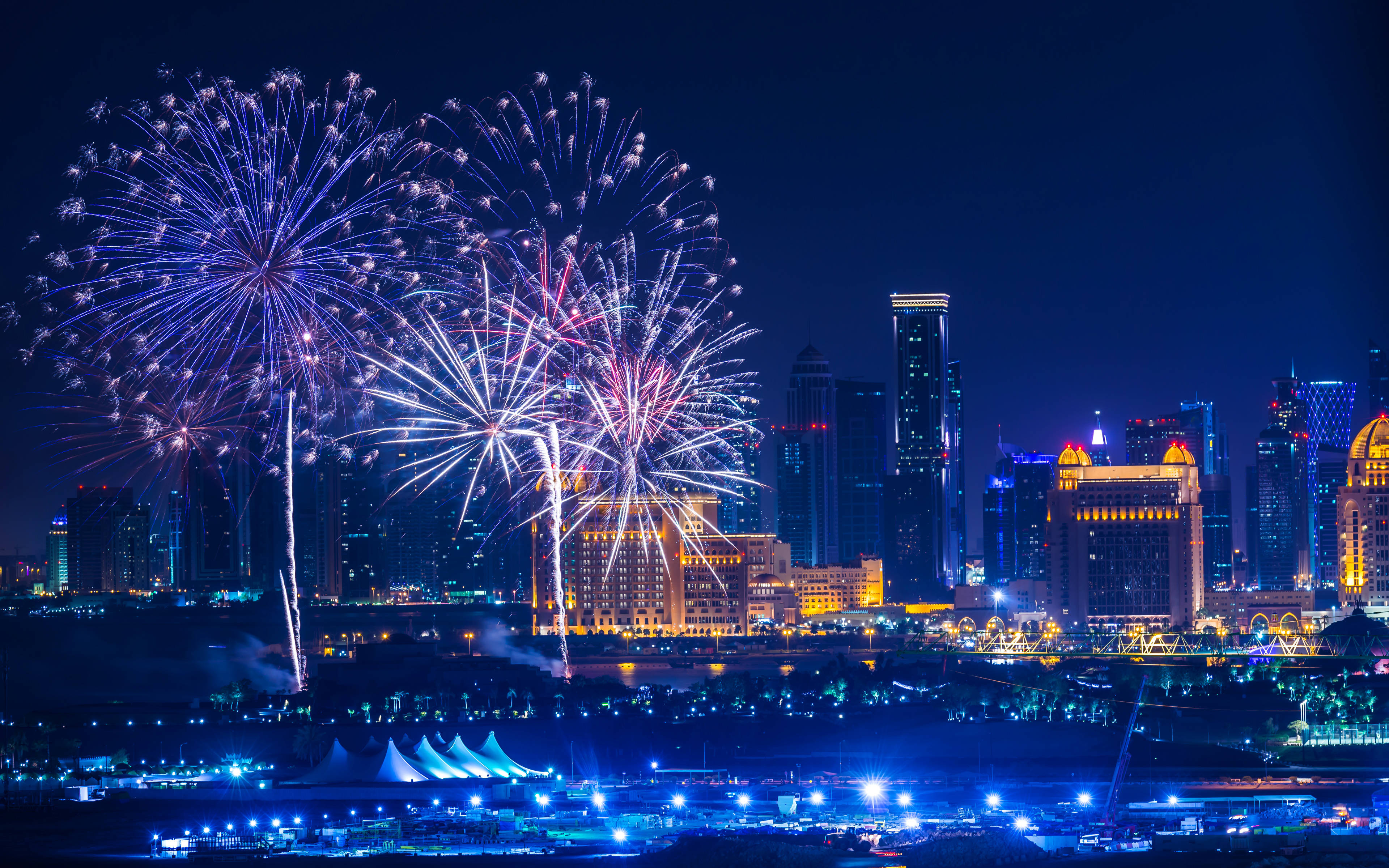 Qatar 桌布 壁紙 — 充滿活力的煙火在夜晚閃爍的城市天際線上爆炸，用藍色和白色的光芒照亮黑暗的天空。