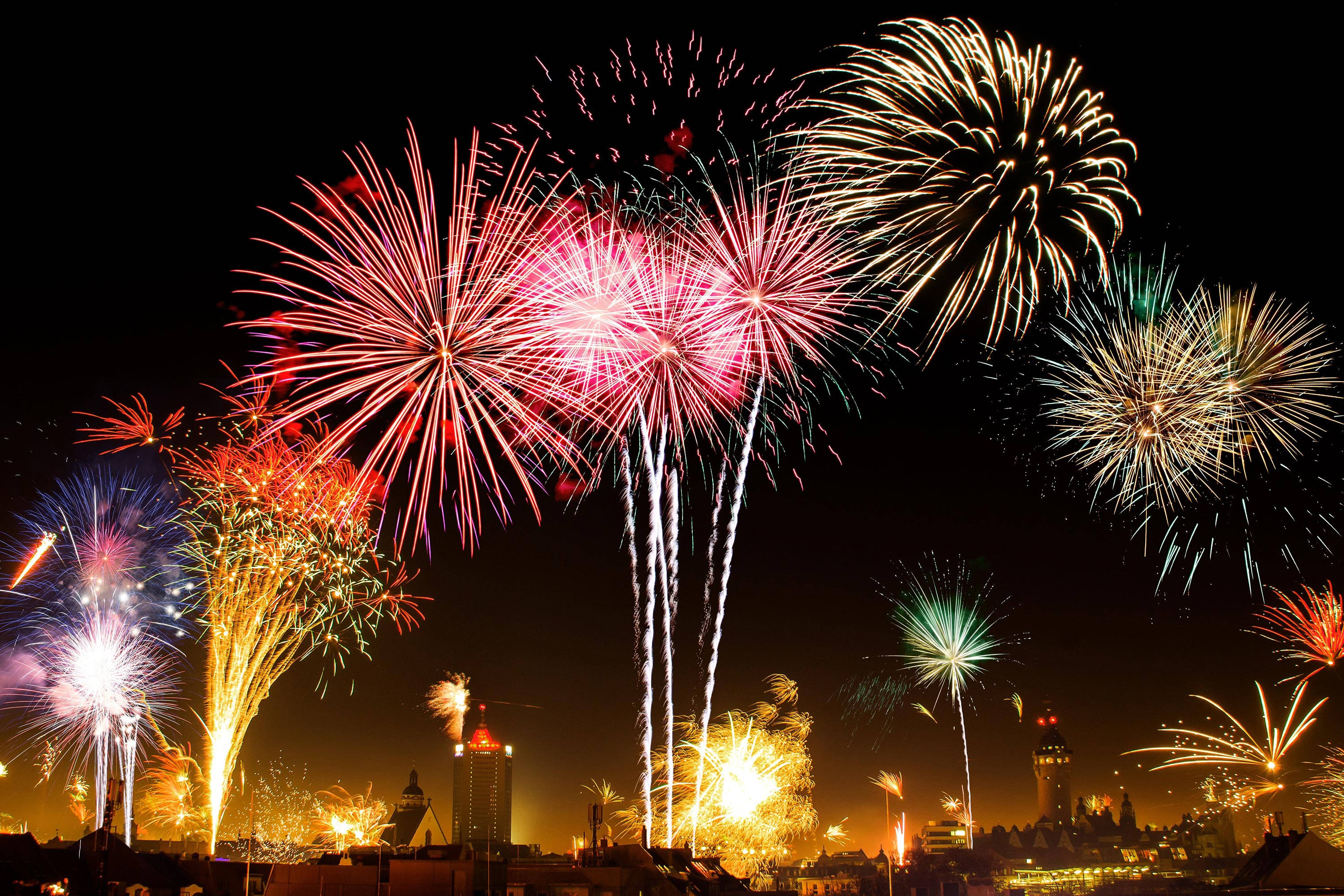Fireworks 桌布 壁紙 — 充滿活力的煙火在夜間城市天際線上綻放出耀眼的光芒，以紅色、金色和藍色色調照亮黑暗的天空。