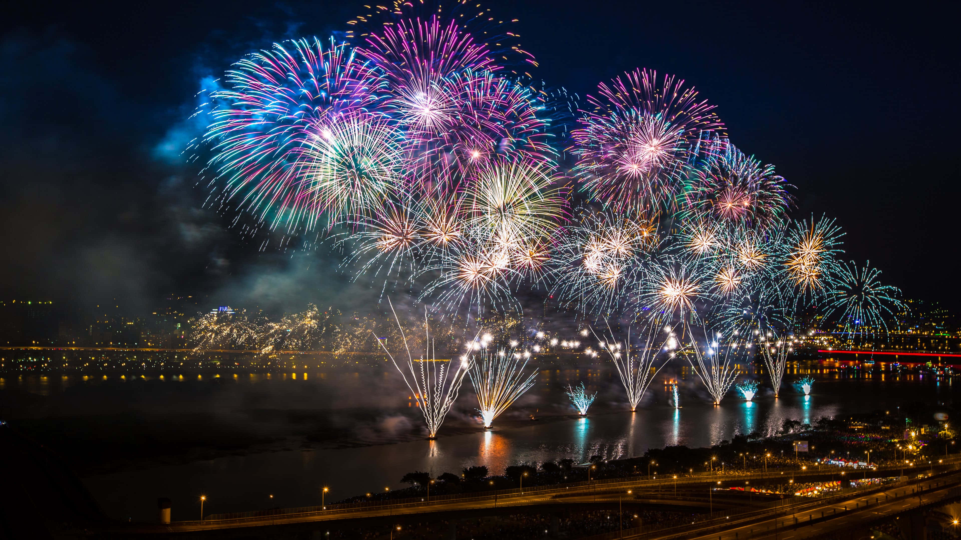 firework 桌布 壁紙 — 充滿活力的煙火在夜間城市天際線倒映在黑暗水面上方綻放出耀眼色彩。