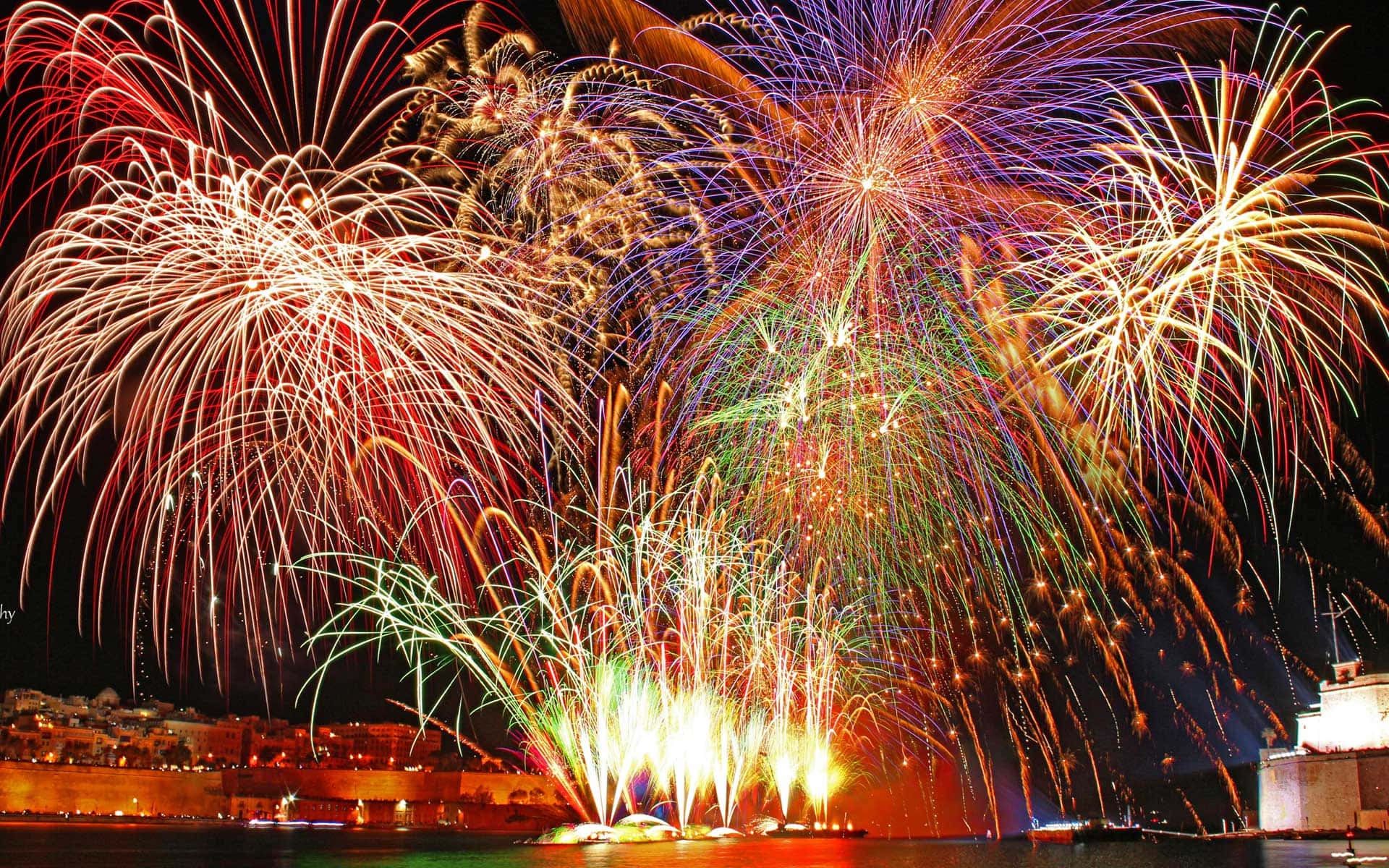 Celebration 桌布 壁紙 — 充滿活力的煙火在夜間於海濱城市上空綻放出耀眼的紅色、綠色和金色光芒。