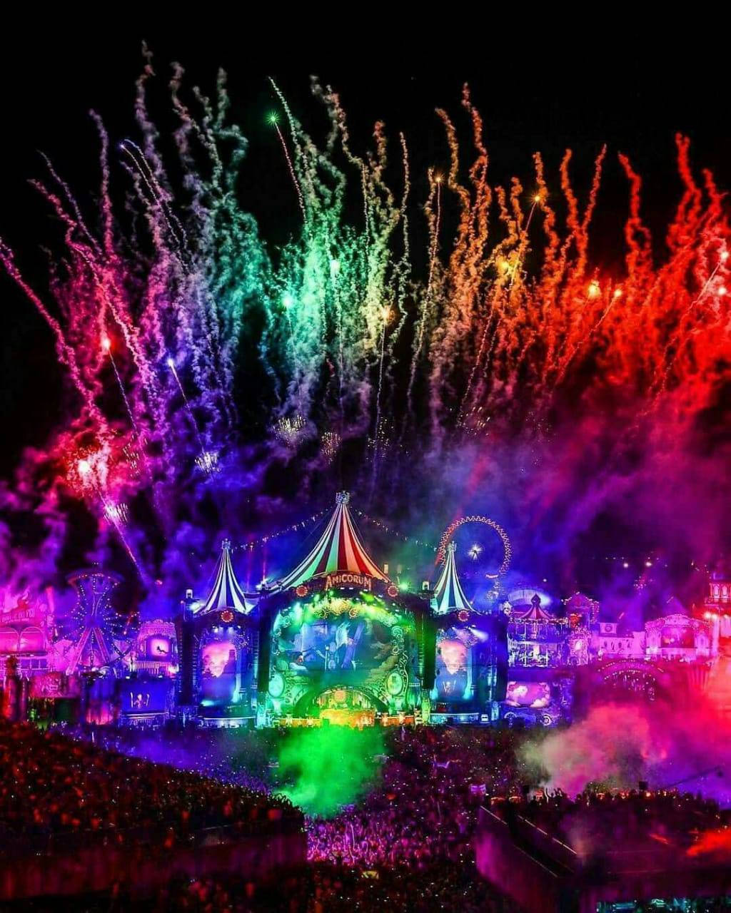 Tomorrowland 桌布 壁紙 — 充滿活力的煙火在彩虹色調中爆炸，在夜晚閃耀的嘉年華舞台上方，照亮下方擠滿的人群。
