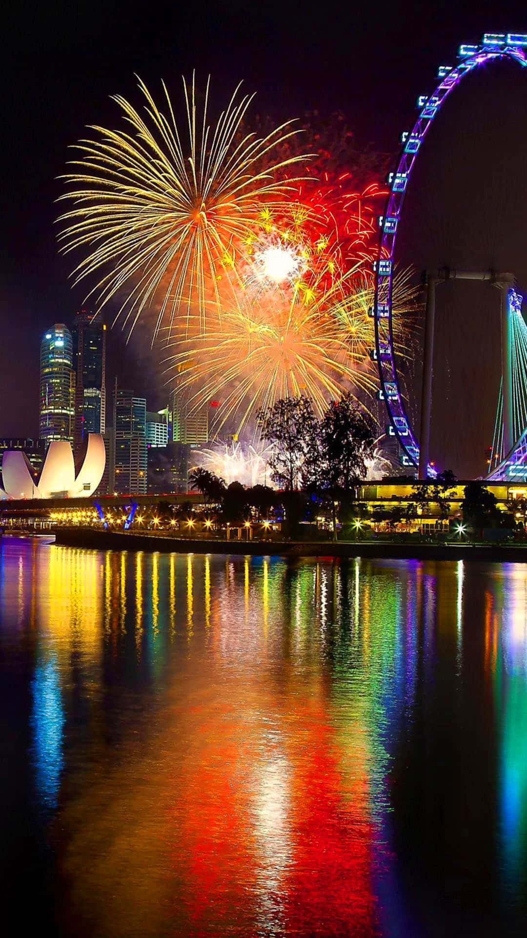 Fireworks 桌布 壁紙 — 充滿活力的煙火在新加坡天際線上綻放，新加坡摩天觀景輪和城市燈光倒映在下方水面上。