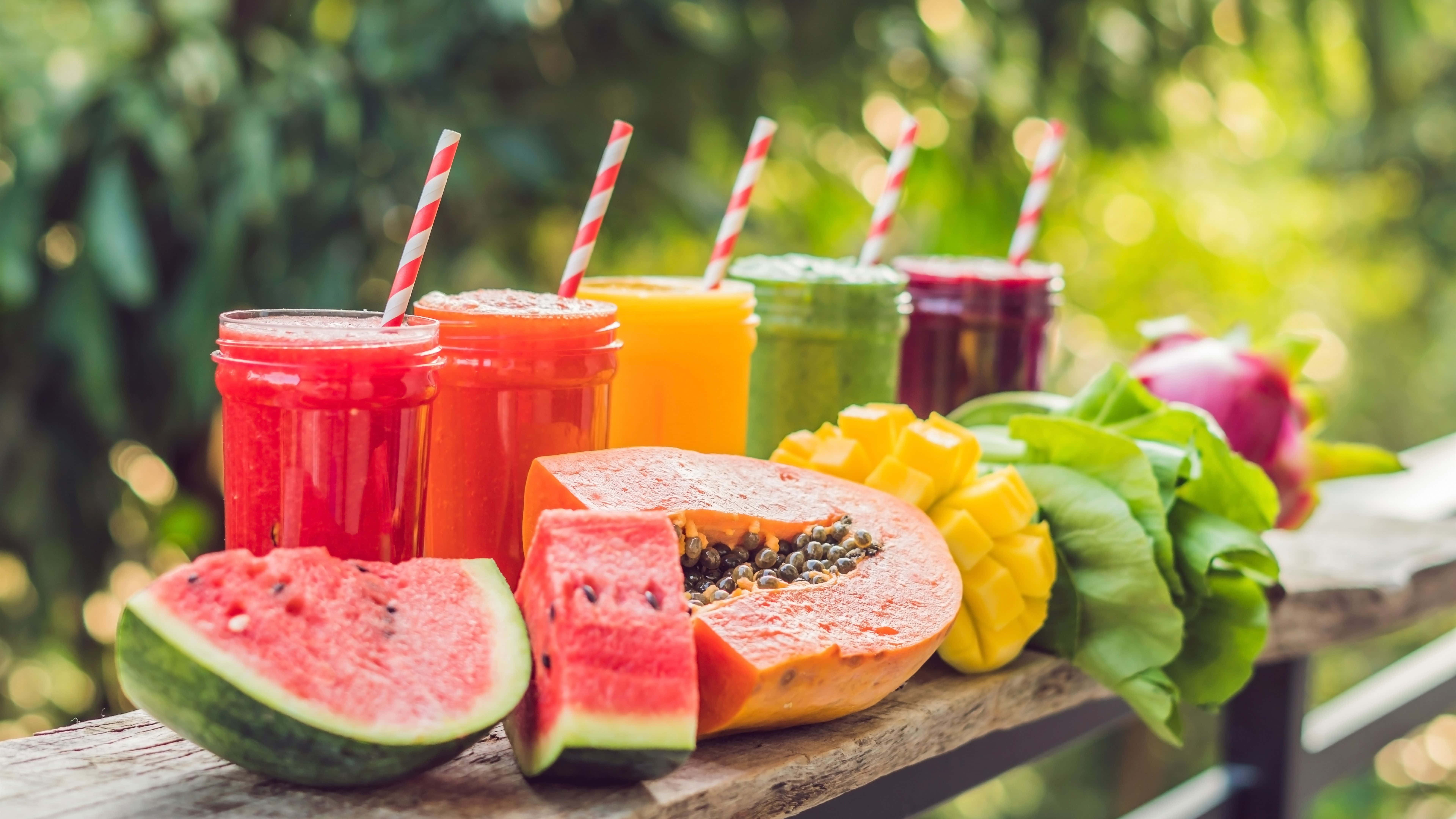 Food And Beverage 桌布 壁紙 — 充滿活力的熱帶水果冰沙裝在玻璃罐中，配上條紋吸管，與新鮮西瓜、木瓜和芒果一起放在木長椅上，斑駁的陽光灑落。