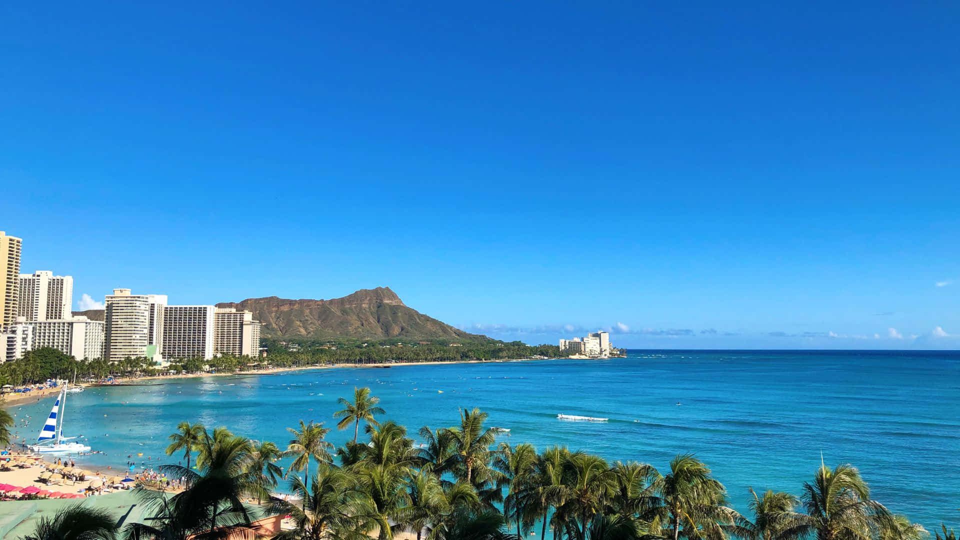 夏威夷 桌布 壁紙 — 充滿活力的熱帶海灘場景，有碧綠的海水、棕櫚樹和一座標誌性的火山，在晴朗的藍天下聳立在現代酒店後面。