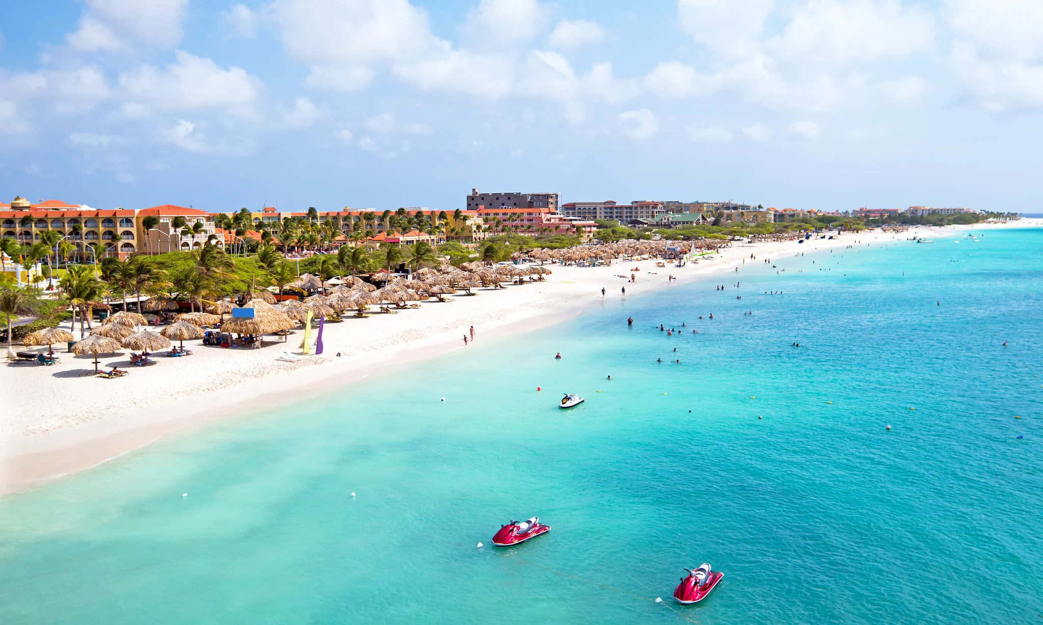 Aruba 桌布 壁紙 — 充滿活力的熱帶海灘鳥瞰圖，碧綠海水、水上摩托車和度假村建築在明亮的藍天下。