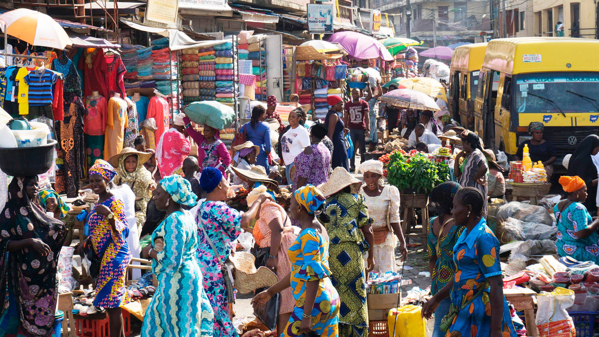 Nigeria 桌布 壁紙 — 充滿活力的熱鬧戶外市場，色彩繽紛的布料、人群和黃色車輛在明亮的日光下。
