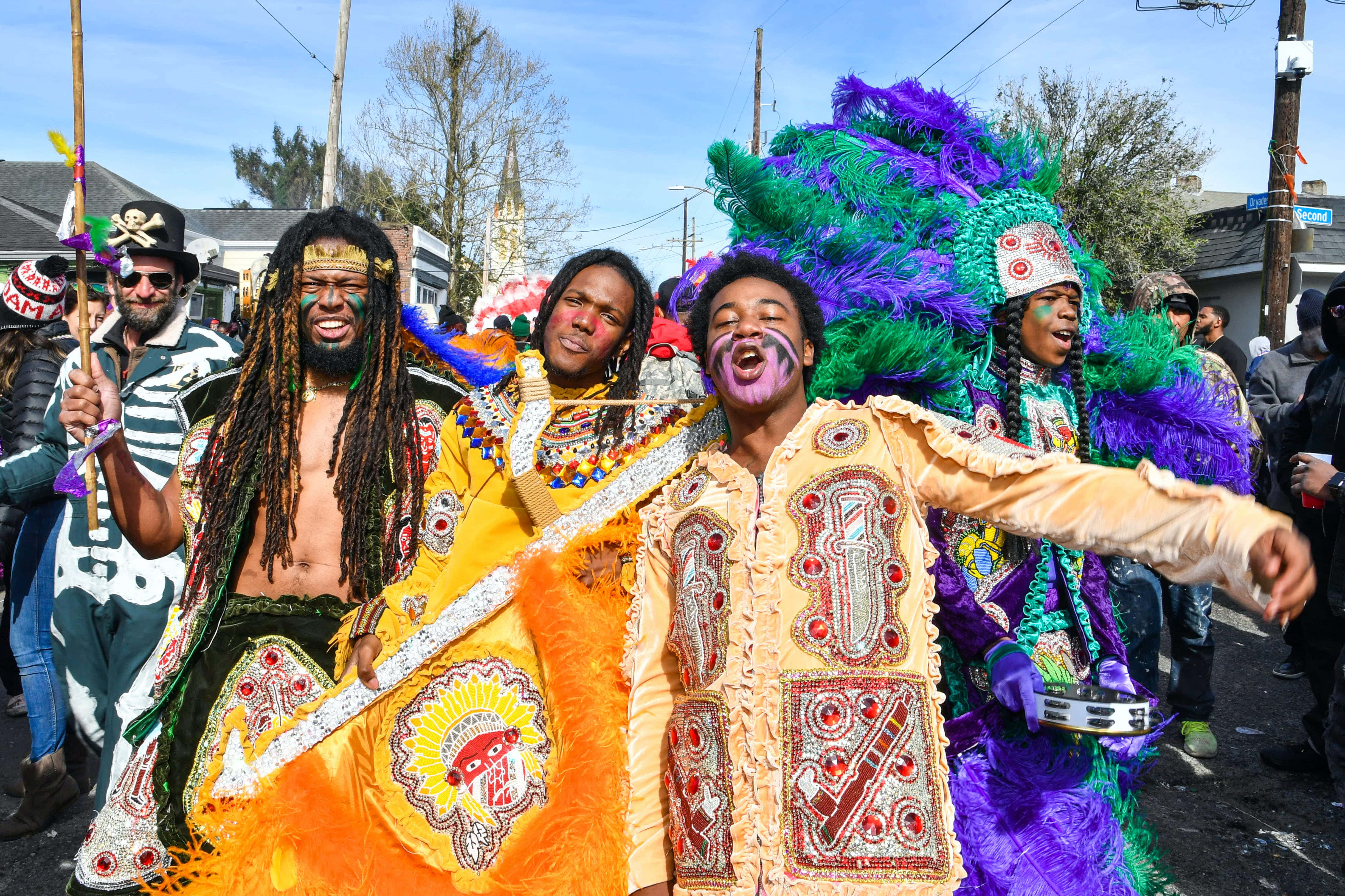 Mardi Gras 桌布 壁紙 — 充滿活力的狂歡節參與者穿著精緻的羽毛服飾和臉部彩繪，在陽光明媚的街道上歡快地跳舞。