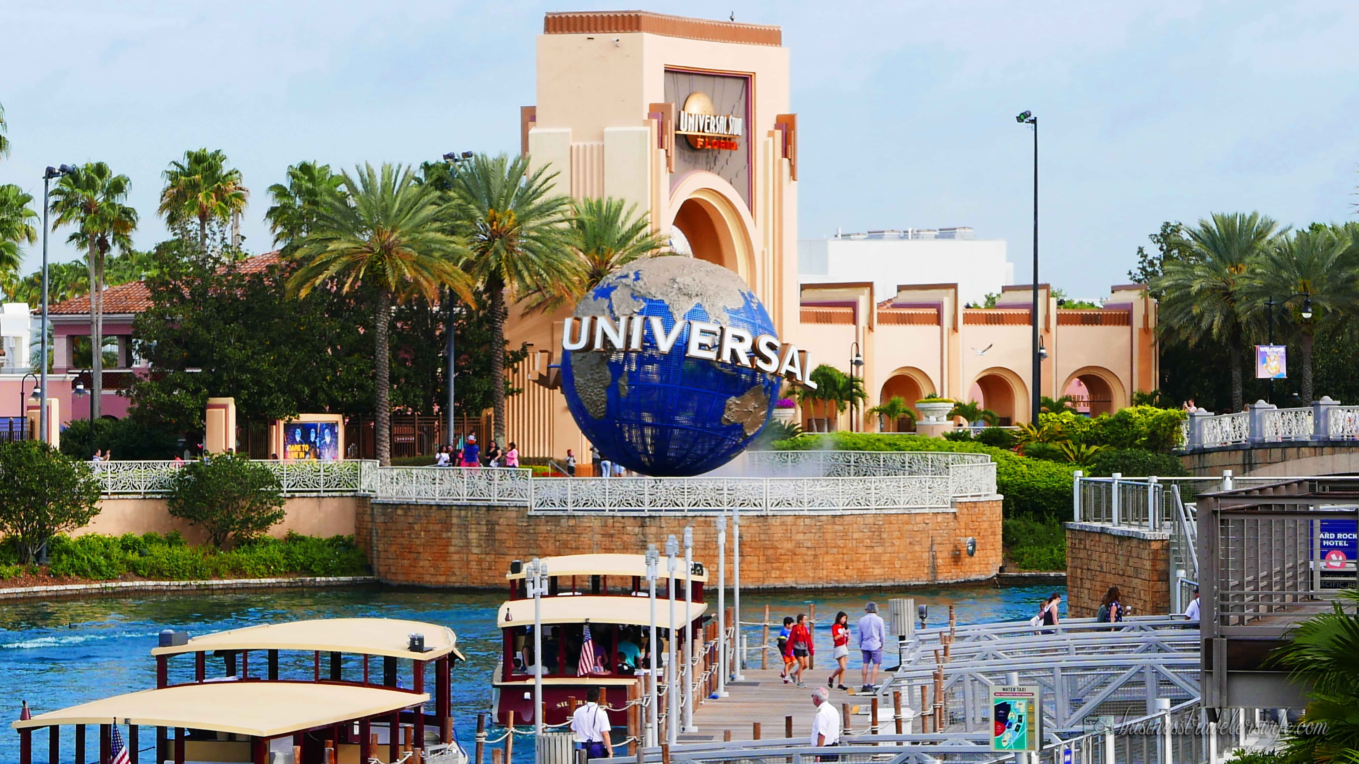 Universal 桌布 壁紙 — 充滿活力的環球影城入口，有一個巨大的藍色地球儀，上面有公園的名稱，周圍環繞著棕櫚樹和船隻，在晴朗的天空下。
