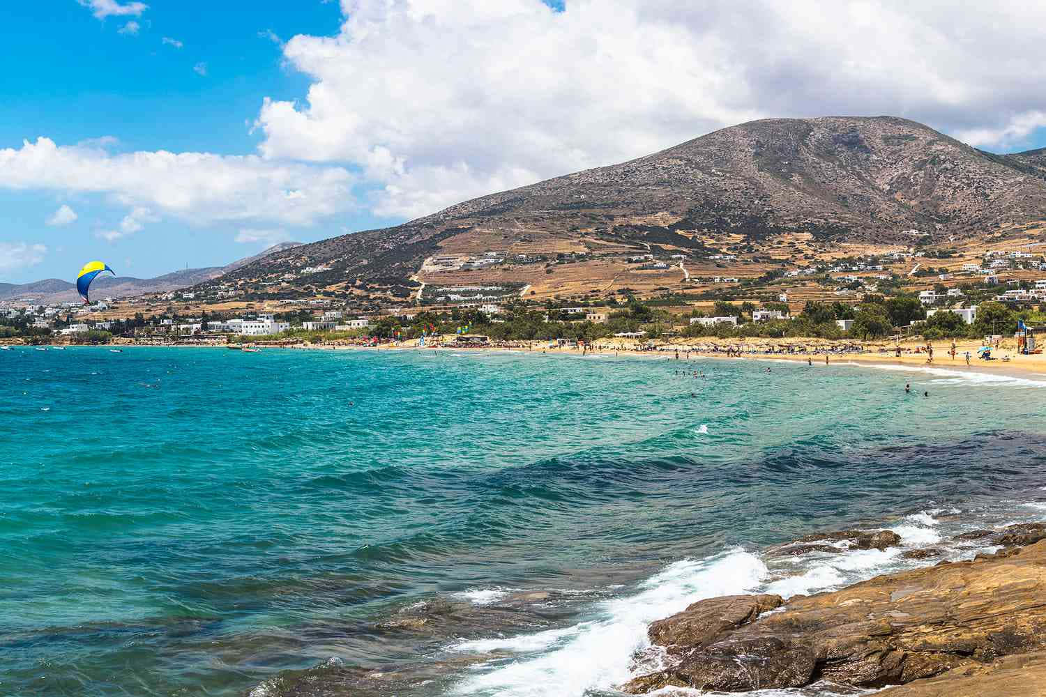 Greek Beach 桌布 壁紙 — 充滿活力的碧綠海水拍打岩石海岸，在明亮的藍天下，點綴著蓬鬆的雲朵，靠近一個陽光明媚的海灘小鎮。