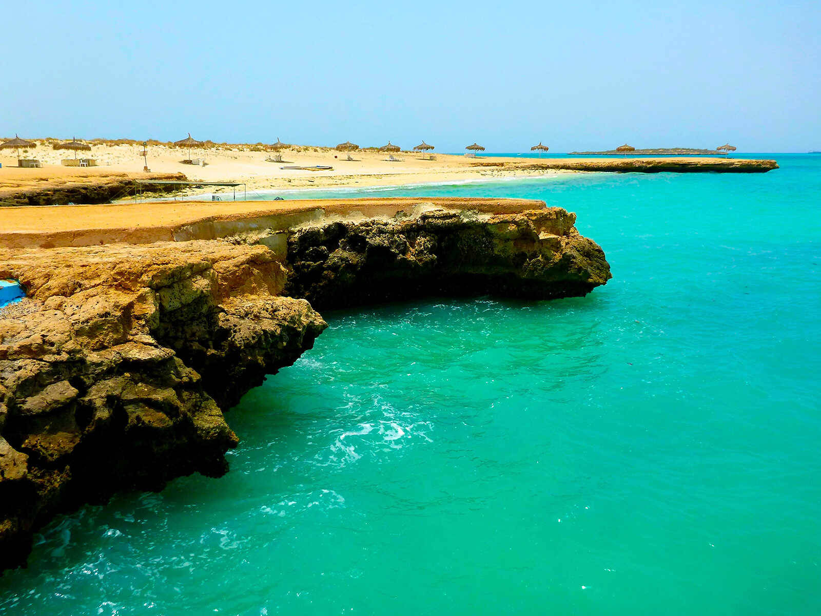 Blue Sky 桌布 壁紙 — 充滿活力的碧綠海水拍打著堅固的金色懸崖，俯瞰著寧靜的沙灘，沙灘上有茅草遮陽傘，天空湛藍清澈。