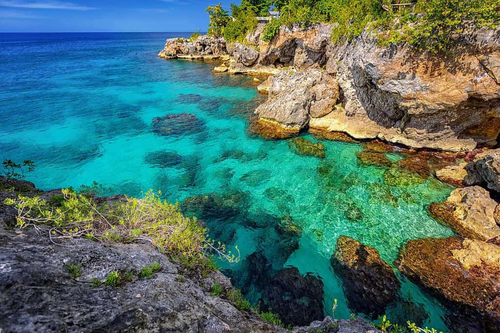 Jamaican Island 桌布 壁紙 — 充滿活力的碧綠海水拍打著崎嶇的懸崖，懸崖上點綴著茂密的綠色植物，在清澈的藍天下。