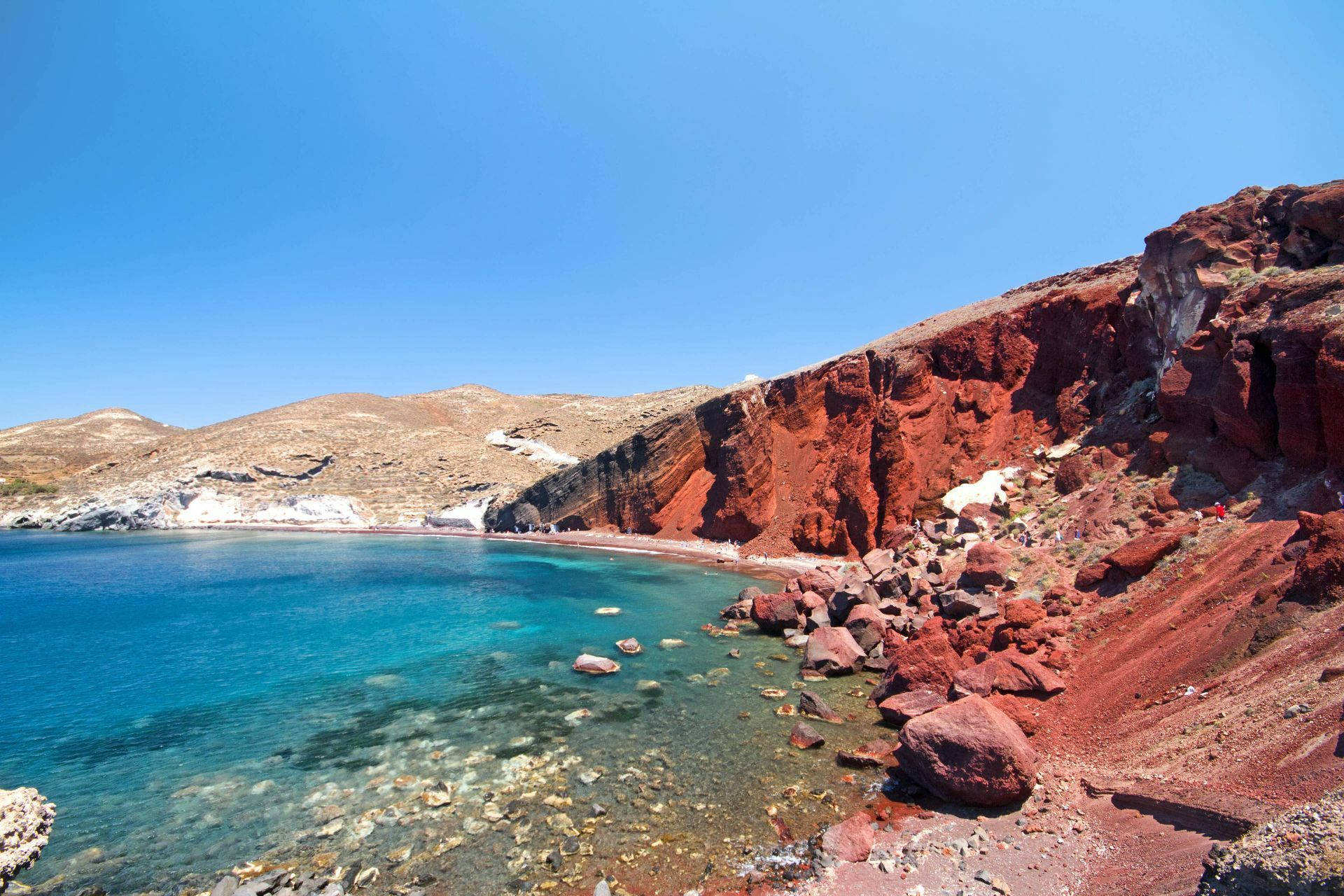 Santorini 桌布 壁紙 — 充滿活力的碧綠海水拍打著引人注目的紅色岩石海岸，上方是清澈的藍天。