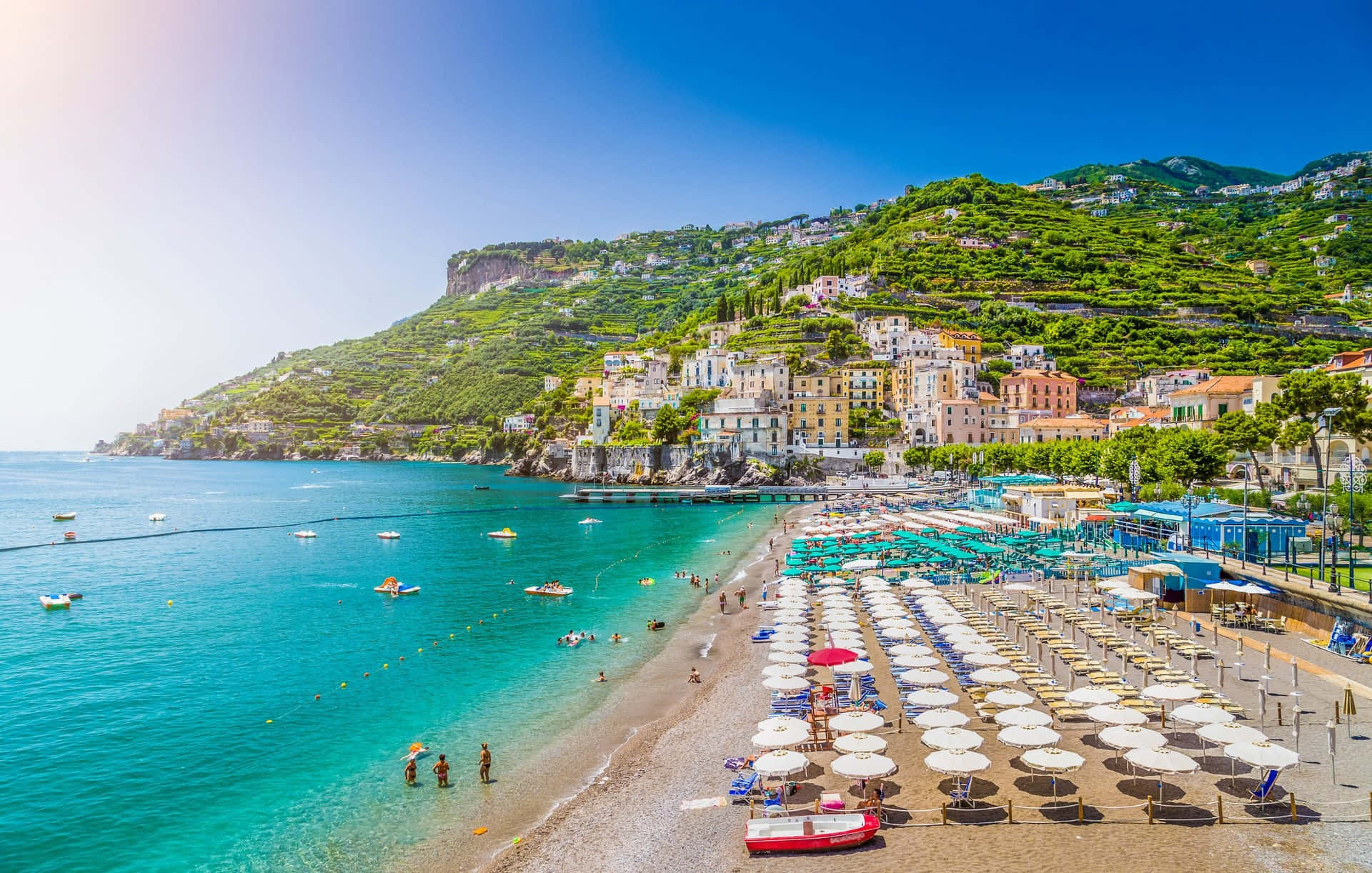 Italian Beach 桌布 壁紙 — 充滿活力的碧綠海水拍打著陽光普照的海灘，海灘上排列著白色遮陽傘和色彩繽紛的懸崖建築，頭頂是清澈的藍天。