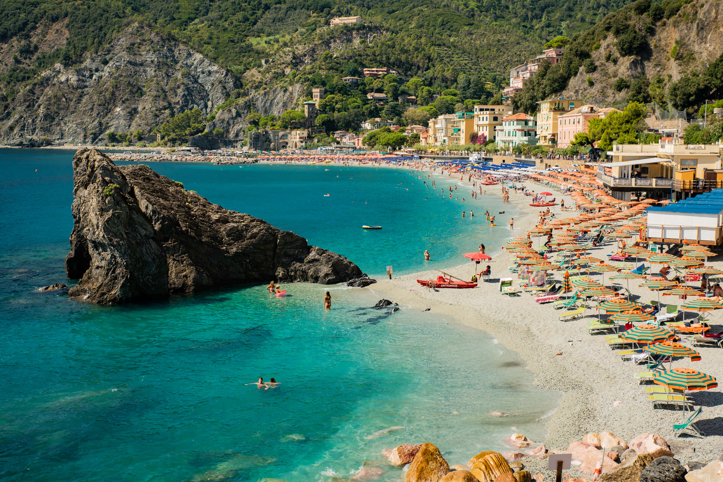Mediterranean Beach 桌布 壁紙 — 充滿活力的碧綠海水拍打著陽光普照的海灘，海灘上擠滿了色彩繽紛的遮陽傘和游泳者，附近有壯觀的岩石突出。