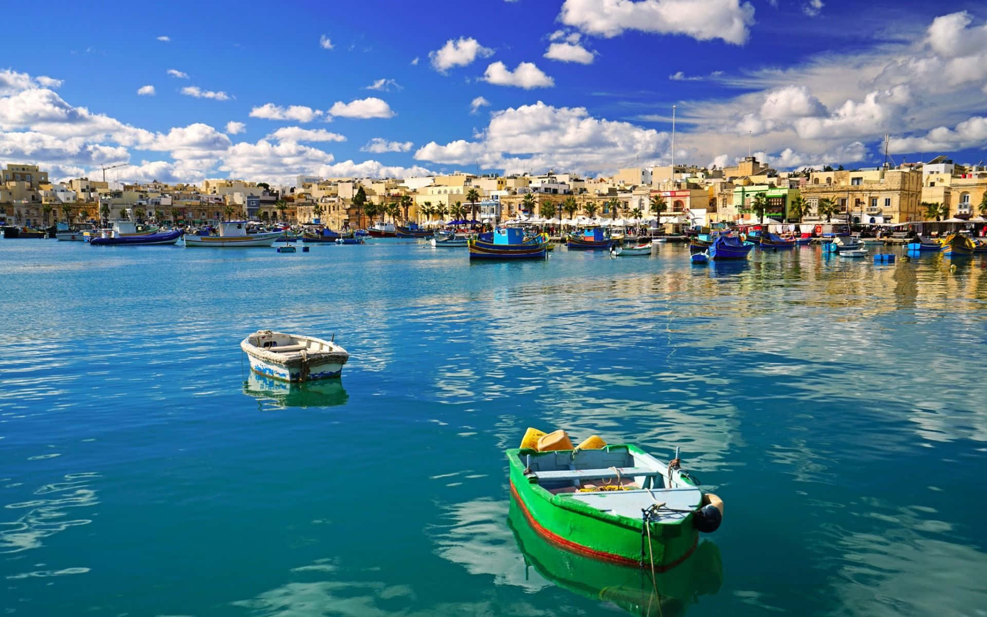 Malta 桌布 壁紙 — 充滿活力的碧綠海水映照著明亮的藍天，色彩繽紛的漁船漂浮在陽光普照的沿海小鎮附近，那裡有陶土屋頂的建築。