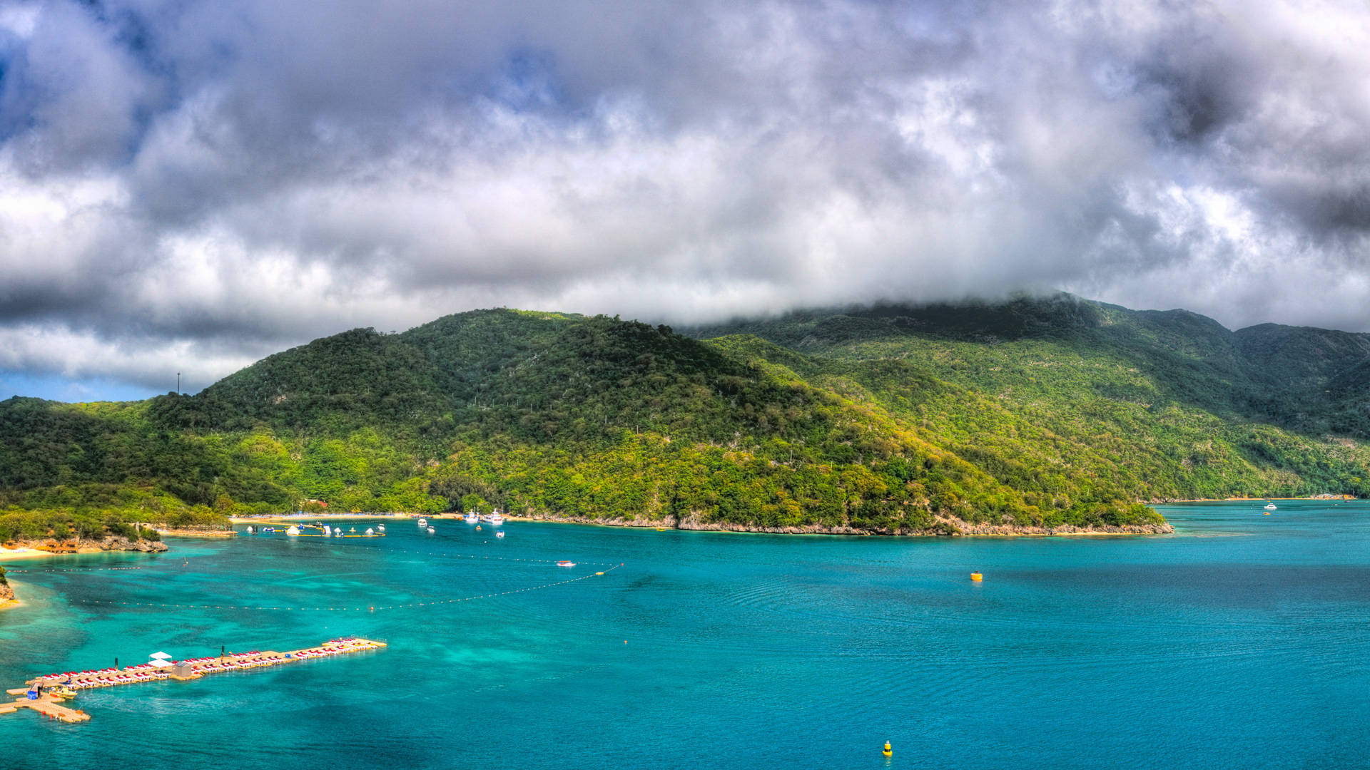 Caribbean 桌布 壁紙 — 充滿活力的碧綠海水環抱著一個鬱鬱蔥蔥的綠色島嶼，天空多雲，戲劇性十足，海灣上點綴著船隻。