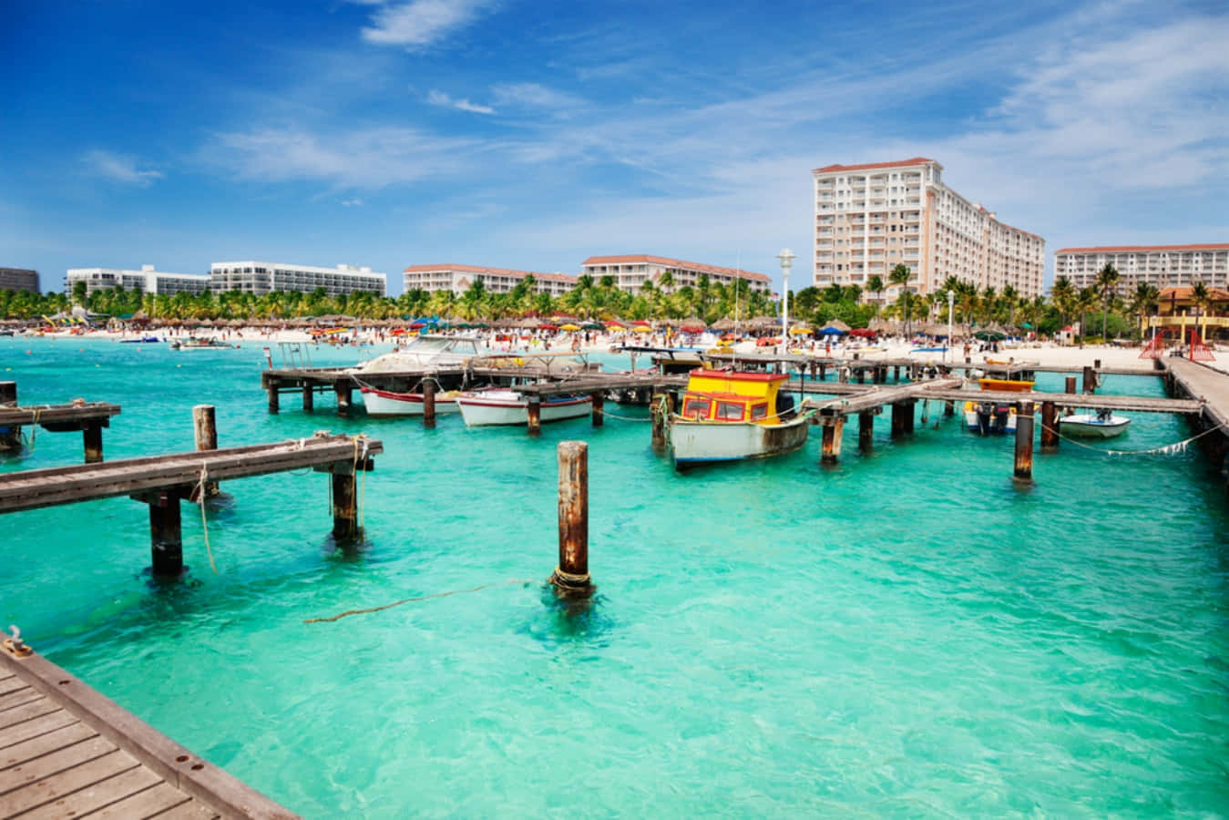 Aruba 桌布 壁紙 — 充滿活力的碧綠海水環繞著風化的木製碼頭，小船停泊在熱鬧的海灘附近，沙灘上棕櫚樹和度假村建築林立，天空湛藍明亮。