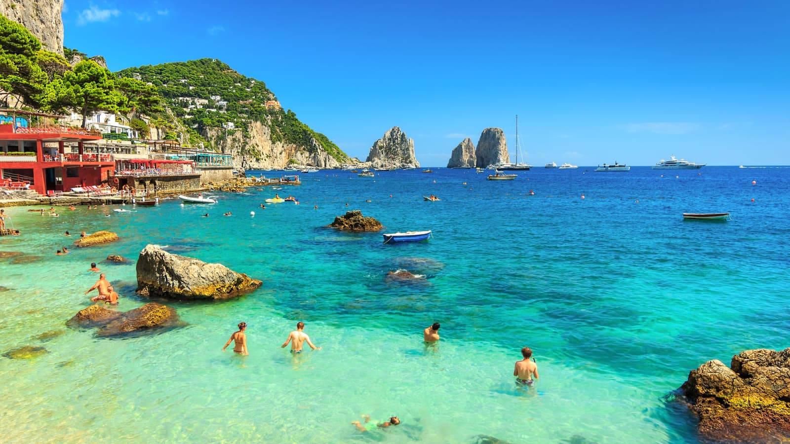 Capri 桌布 壁紙 — 充滿活力的碧綠海水邀請泳客靠近岩石海岸，懸崖邊的建築物和標誌性的法拉廖尼岩石在清澈的藍天下。