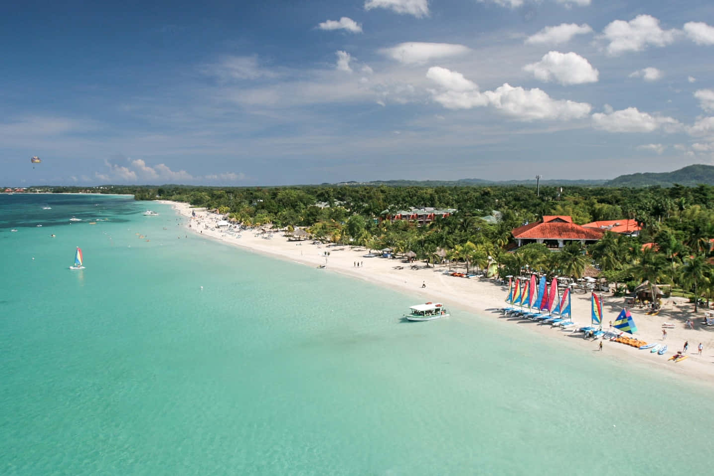 Jamaican Island 桌布 壁紙 — 充滿活力的碧綠海灘鳥瞰圖，白色沙灘上種滿棕櫚樹，色彩繽紛的帆船在明亮的藍天下航行。