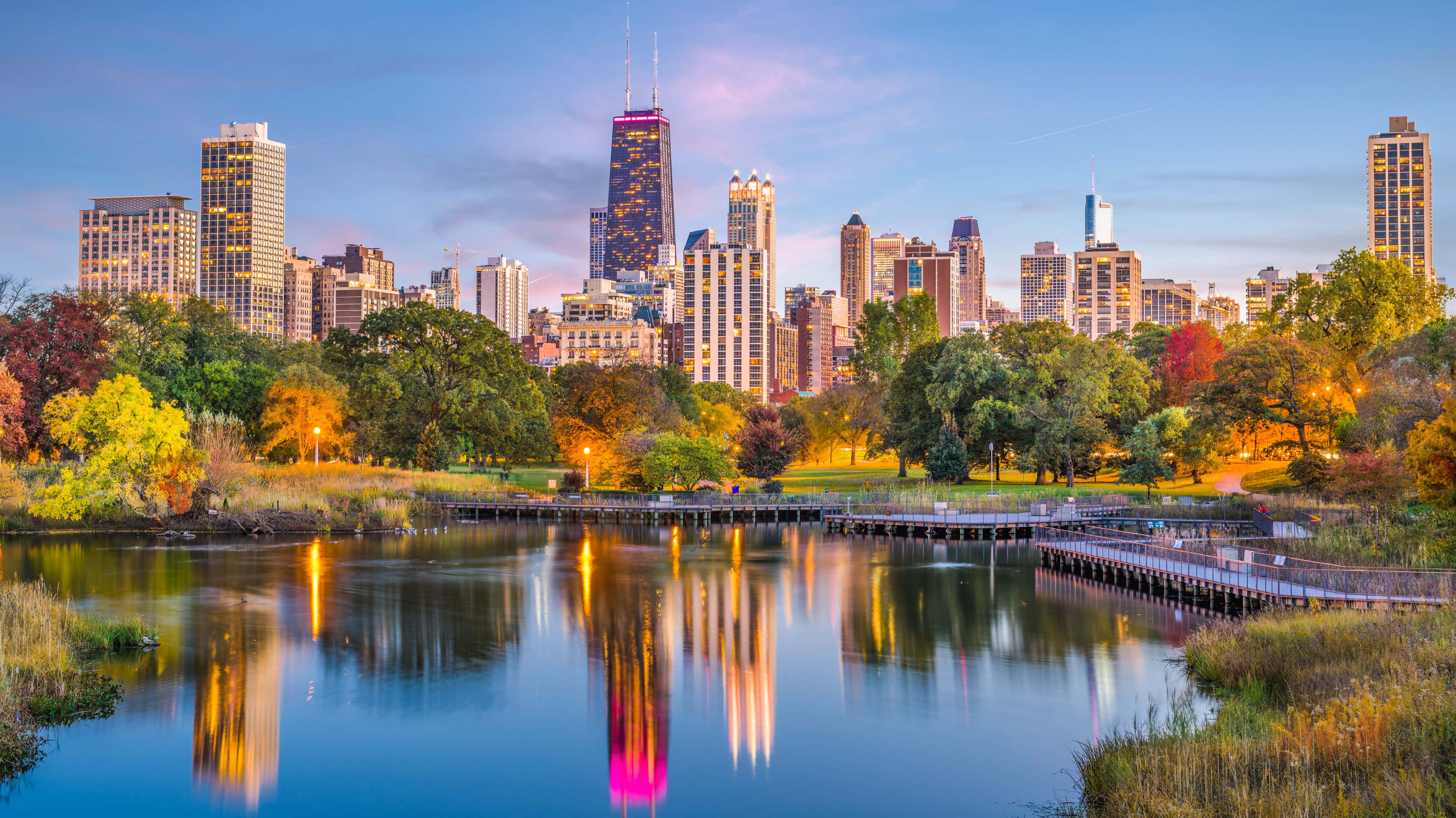 4K Ultra HD Chicago 桌布 壁紙 — 充滿活力的秋日城市景觀，摩天大樓倒映在平靜的湖面上，周圍環繞著色彩繽紛的樹葉，夕陽西下。