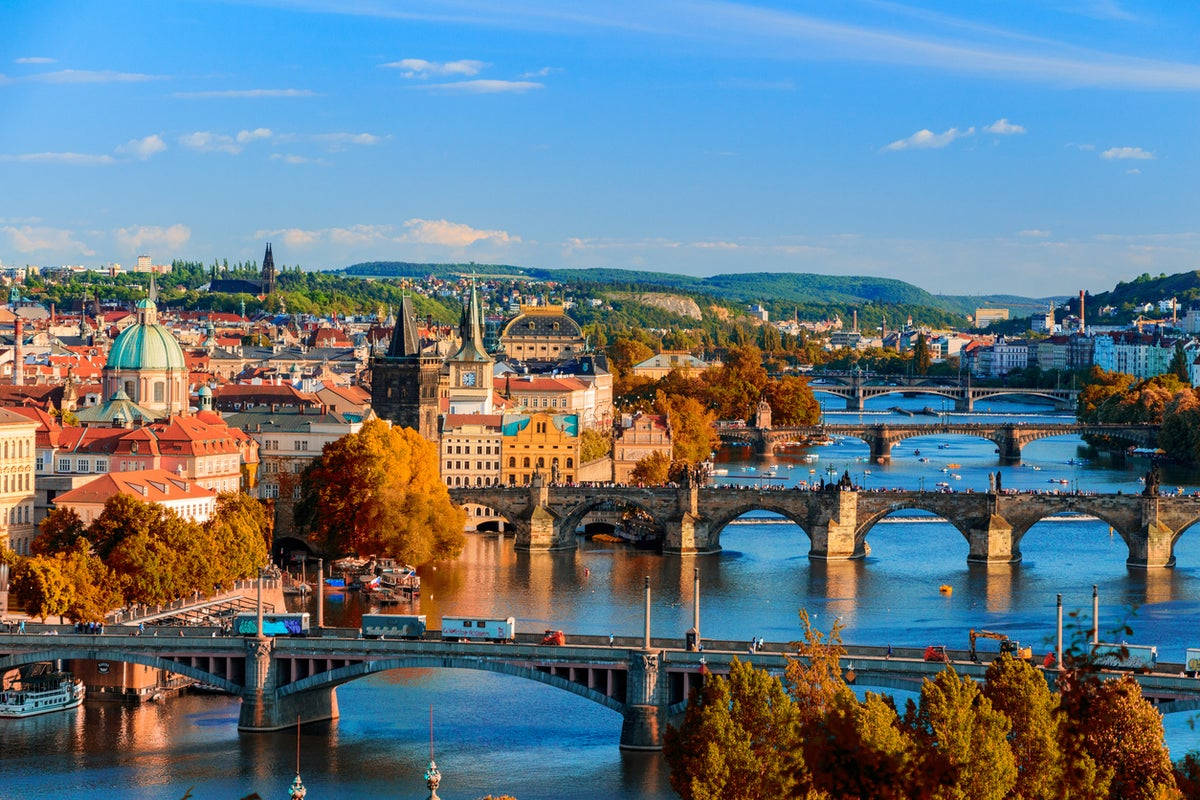Europe 桌布 壁紙 — 充滿活力的秋日城市景觀，歷史悠久的橋樑橫跨河流，四周環繞著色彩繽紛的樹葉和紅瓦屋頂，在清澈的藍天下。