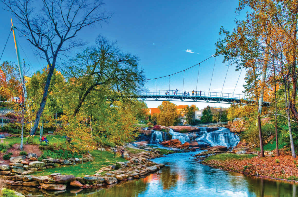 Fall Bridge 桌布 壁紙 — 充滿活力的秋日景色，一座吊橋橫跨層疊的河流，周圍是色彩繽紛的樹木，天空清澈湛藍。