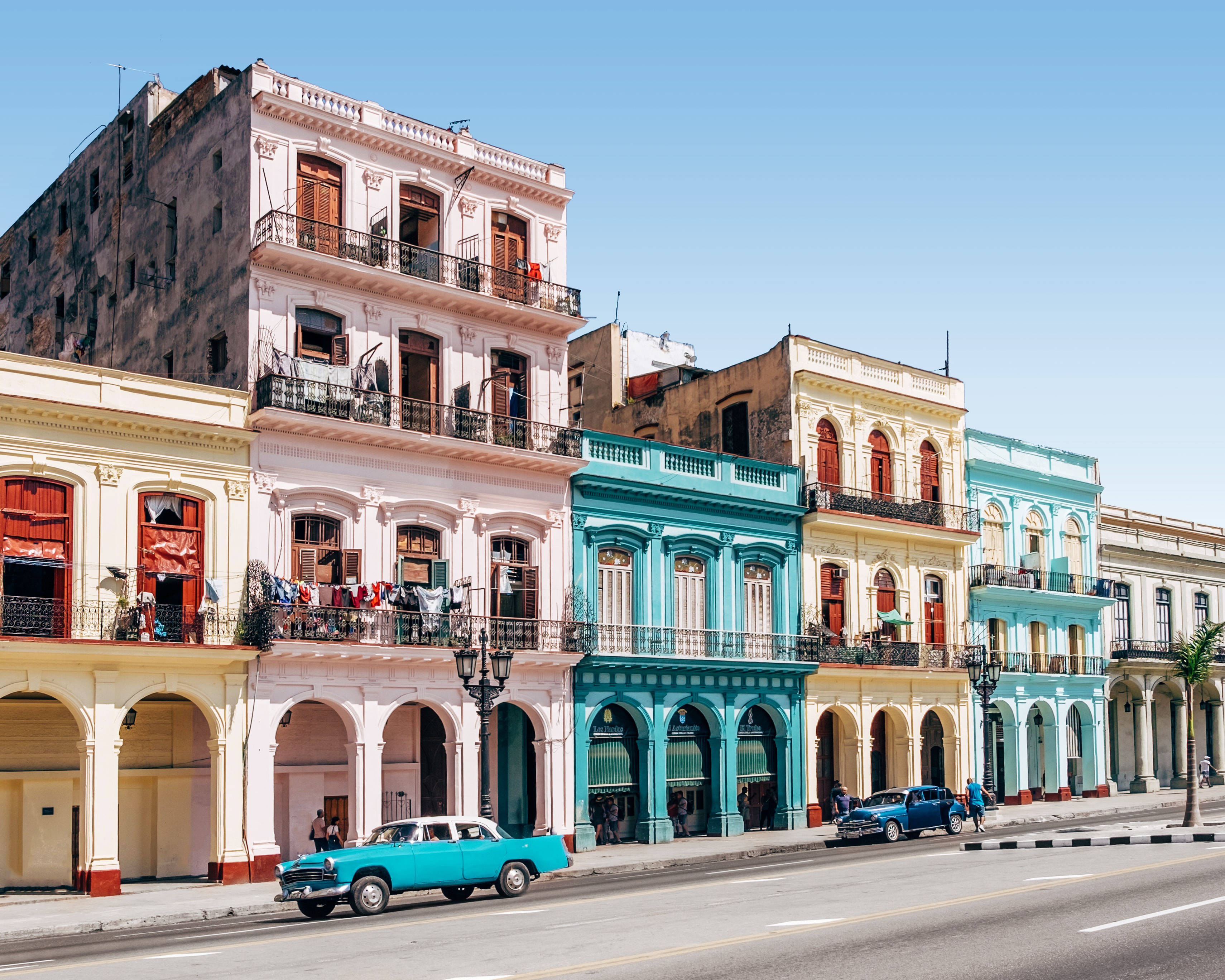 Cuba 桌布 壁紙 — 充滿活力的粉彩殖民地建築排列在陽光普照的街道上，經典美國汽車沿路邊停放。