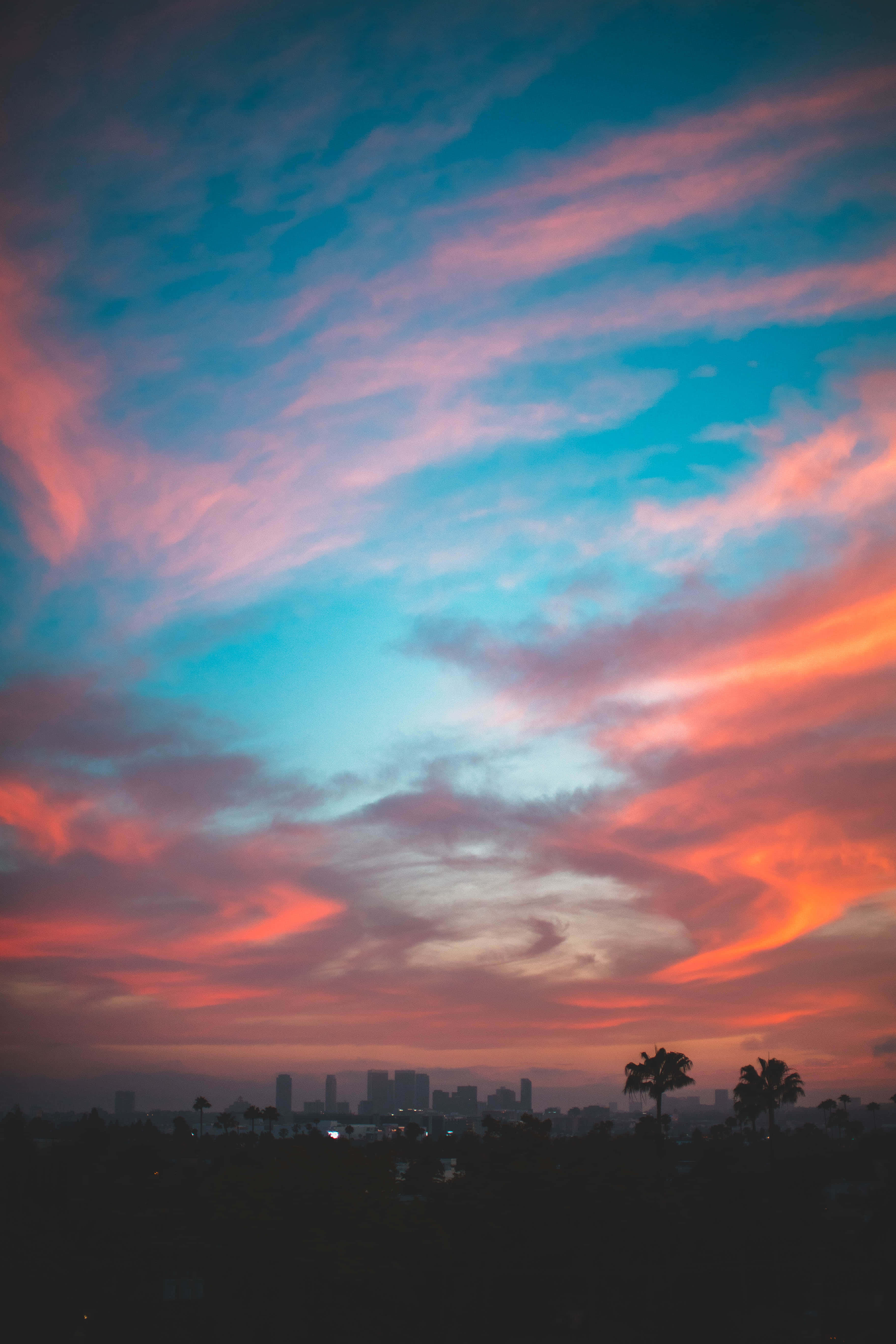 Clouds Aesthetics 桌布 壁紙 — 充滿活力的粉紅色、橙色和藍綠色雲彩在暮色天空中延伸，映襯著剪影般的城市景觀和棕櫚樹。