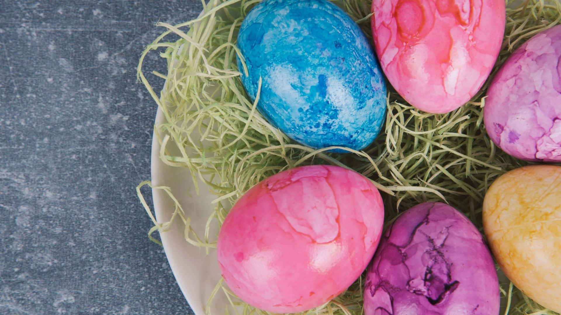 Easter Egg 桌布 壁紙 — 充滿活力的粉紅色、藍色和紫色大理石紋復活蛋，放在白色盤子上的綠色稻草中，背景為灰色紋理。