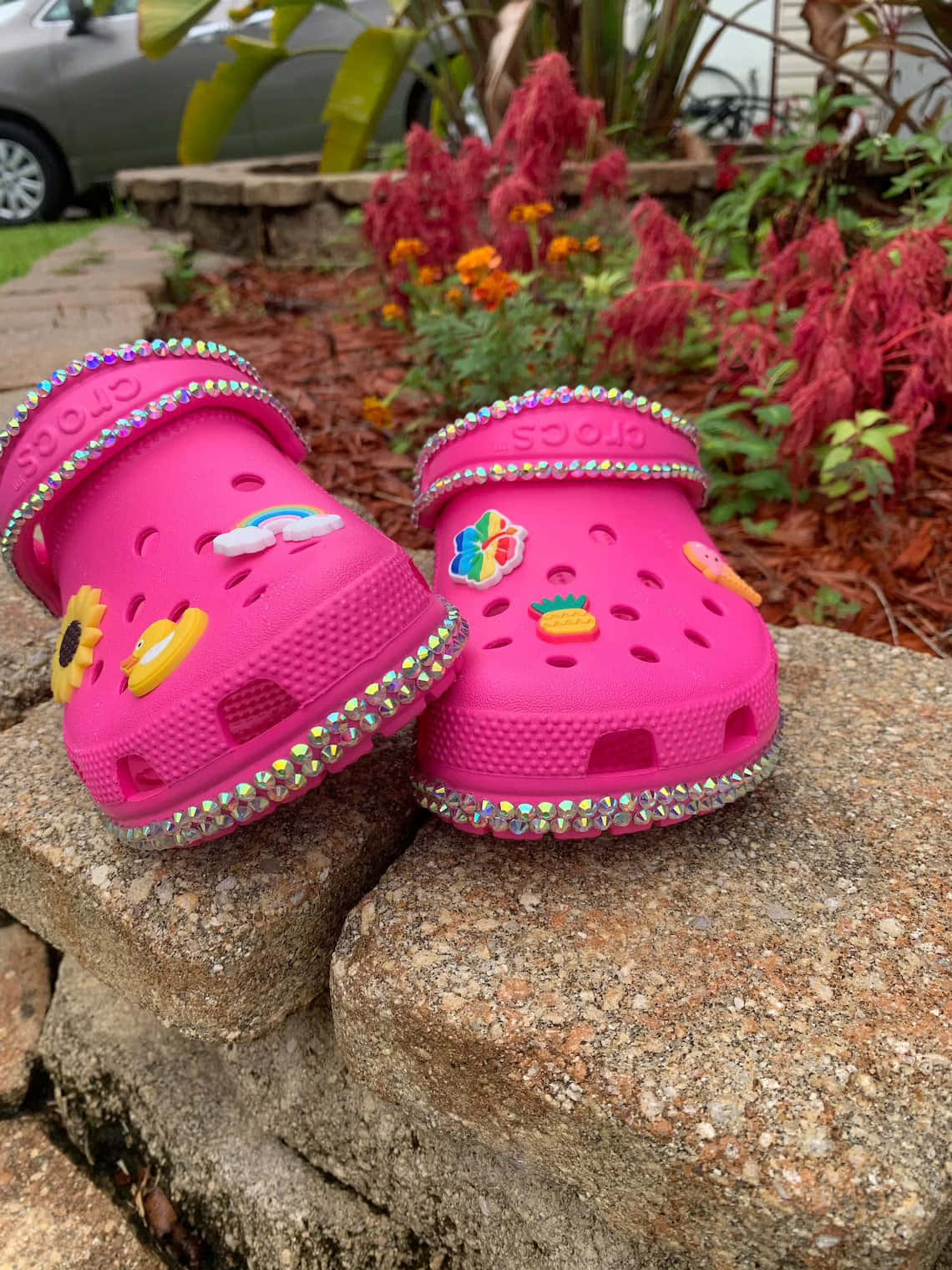 Crocs 桌布 壁紙 — 充滿活力的粉紅色Crocs，飾有水鑽和彩色吊飾，放在石牆上，旁邊有盛開的紅色和橙色花朵。