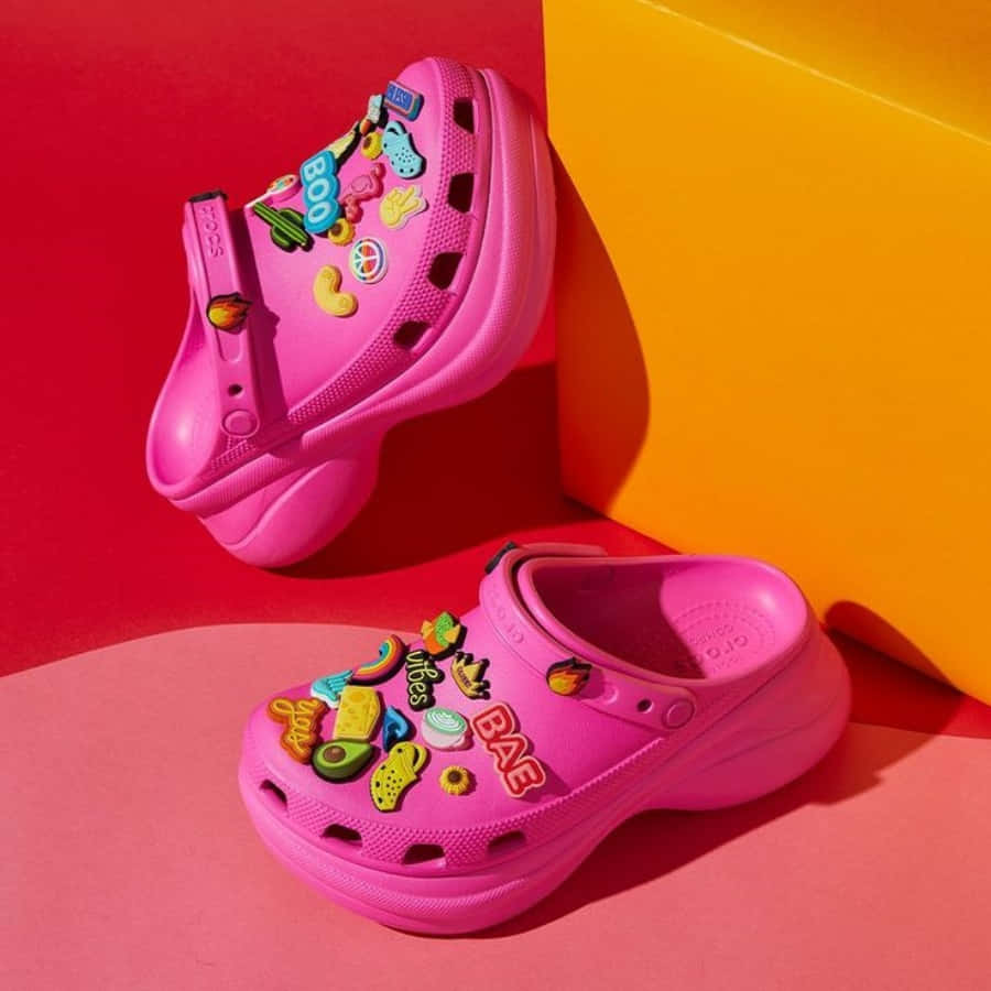 Crocs 桌布 壁紙 — 充滿活力的粉紅色Crocs木屐，覆蓋著色彩繽紛的飾物，在明亮的攝影棚燈光下靠著大膽的紅色和黃色幾何形狀塊。