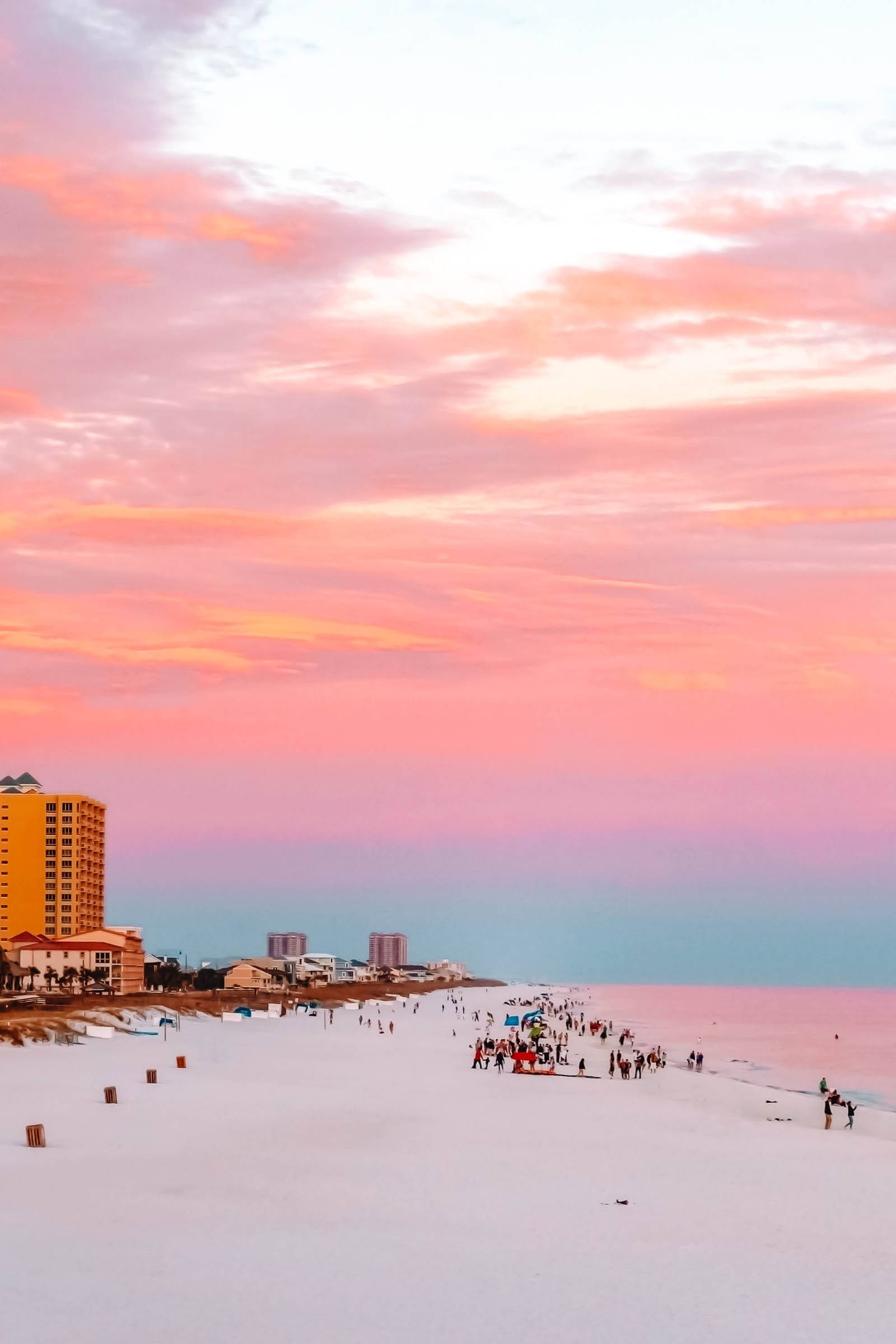 Beautiful Beach 桌布 壁紙 — 充滿活力的粉紅色和橙色夕陽，描繪在擁擠的白色沙灘和遠處飯店的天空上。