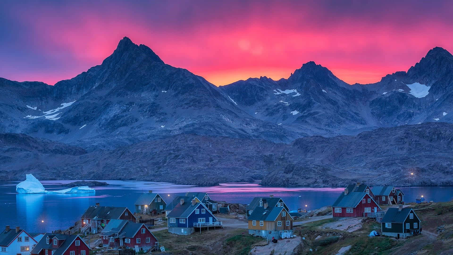 Greenland 桌布 壁紙 — 充滿活力的粉紅色和紫色日落照亮了崎嶇的山脈和寧靜峽灣旁色彩繽紛的房屋，峽灣中漂浮著冰山。