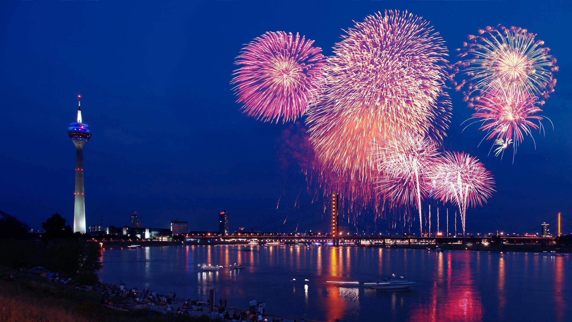 Fireworks 桌布 壁紙 — 充滿活力的粉紅色和金色煙火在寧靜河流中倒映的城市天際線上爆炸。