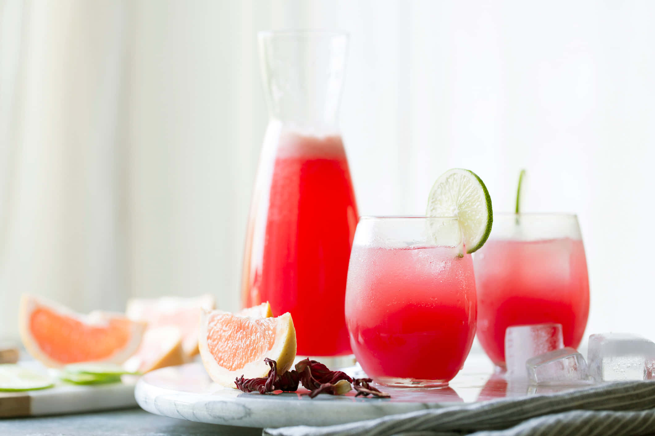 Beautiful Drink 桌布 壁紙 — 充滿活力的粉紅色木槿花飲料，配上青檸裝飾和冰塊，盛在玻璃杯中，旁邊有切片葡萄柚和一個玻璃壺。