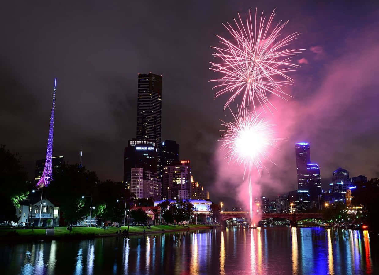 New Years Eve 桌布 壁紙 — 充滿活力的粉紅色煙火在夜間城市天際線上爆炸，倒映在下方平靜的河流中。