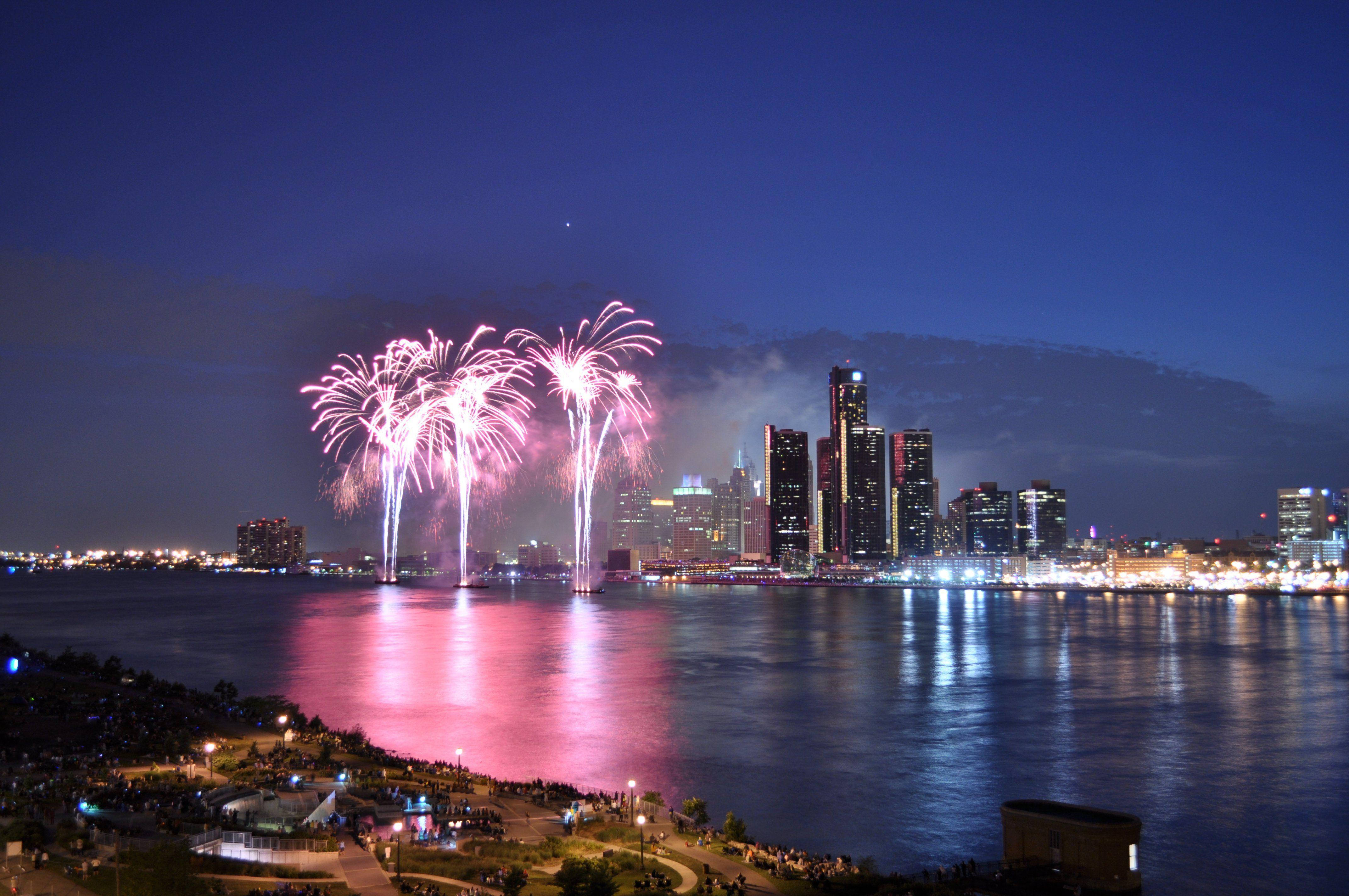 detroit 桌布 壁紙 — 充滿活力的粉紅色煙火在夜間平靜水面上倒映的城市天際線上空爆炸。