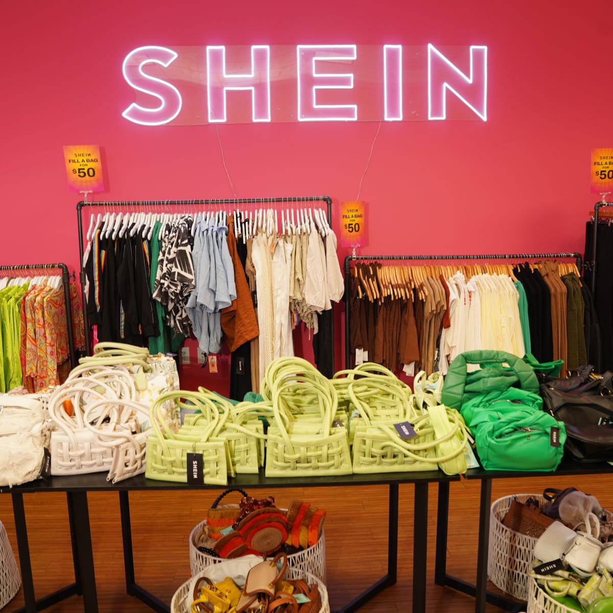 Shein 桌布 壁紙 — 充滿活力的粉紅色牆壁，上面有發光的SHEIN霓虹燈標誌，下方是色彩繽紛的衣服架和萊姆綠色編織手提包。