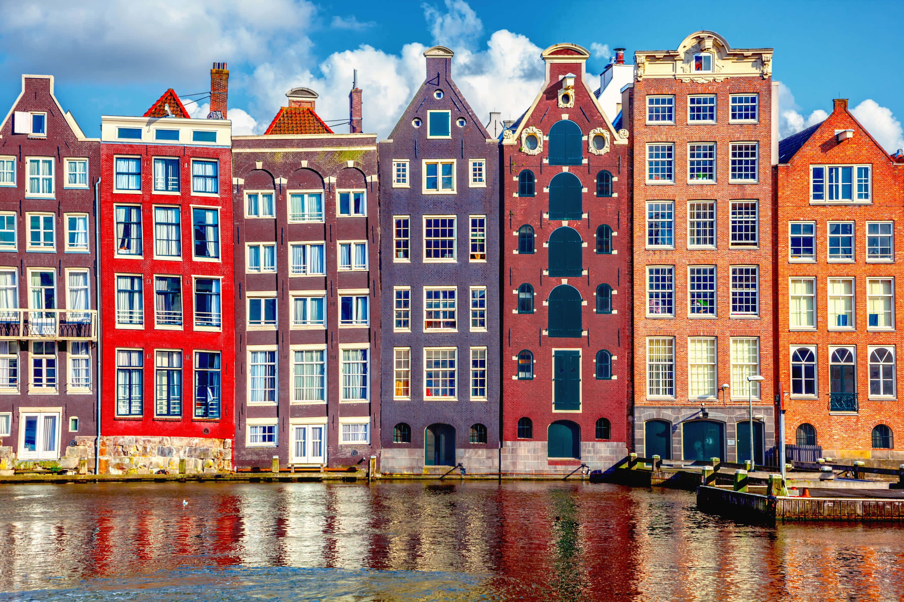 Netherlands 桌布 壁紙 — 充滿活力的紅磚運河房屋，帶有山牆屋頂，在平靜的水面上倒映，天空湛藍，白雲朵朵。