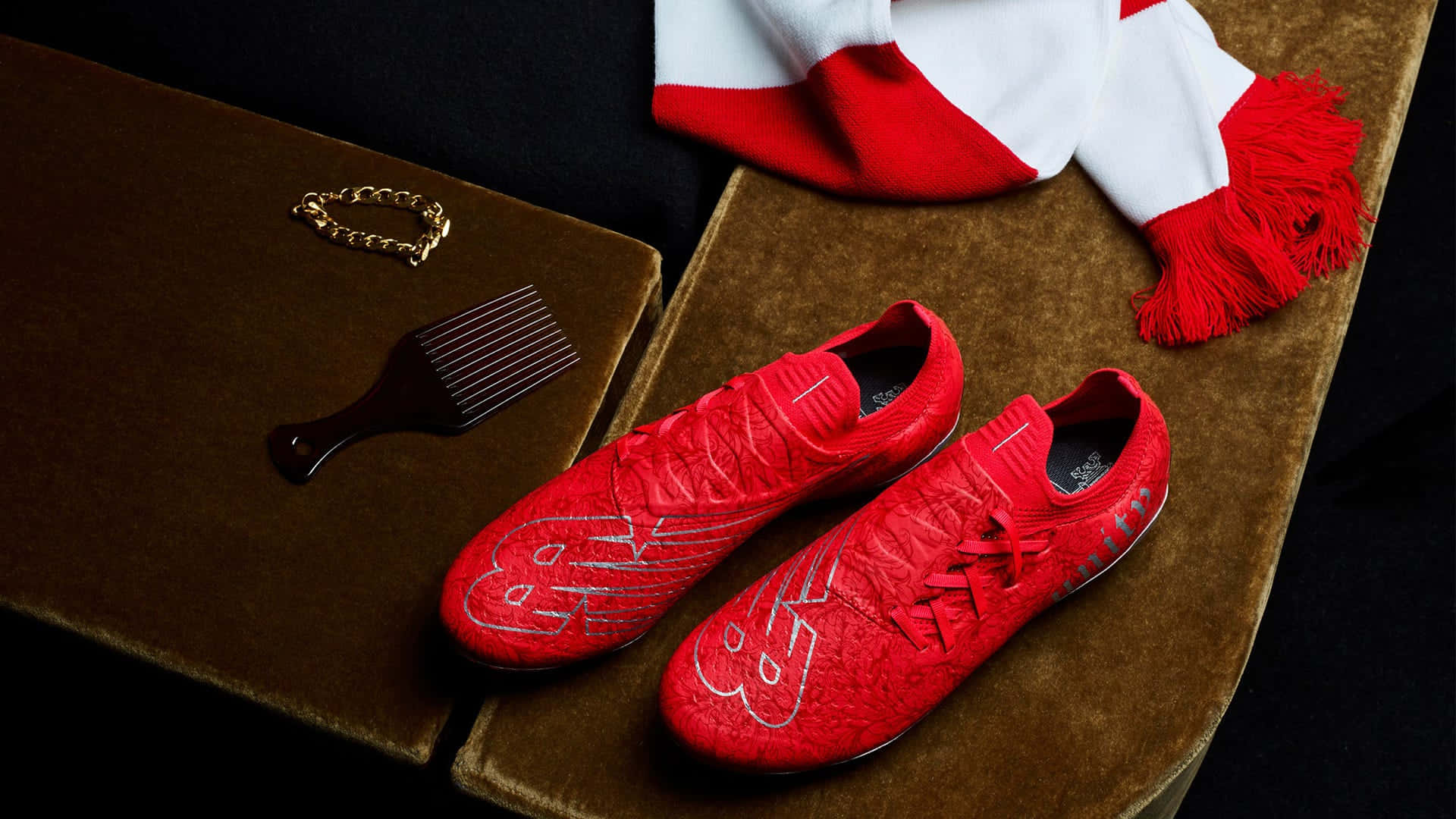 New Balance 桌布 壁紙 — 充滿活力的紅色足球鞋，帶有銀色裝飾，放置在棕色天鵝絨表面上，旁邊有相配的圍巾和護理工具。
