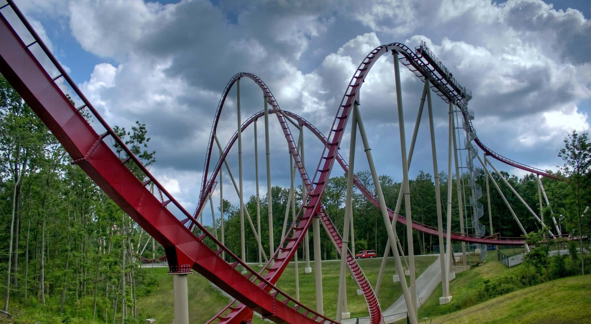 Roller Coaster 桌布 壁紙 — 充滿活力的紅色雲霄飛車在戲劇性的多雲天空下扭轉和環繞，周圍環繞著茂密的綠樹。