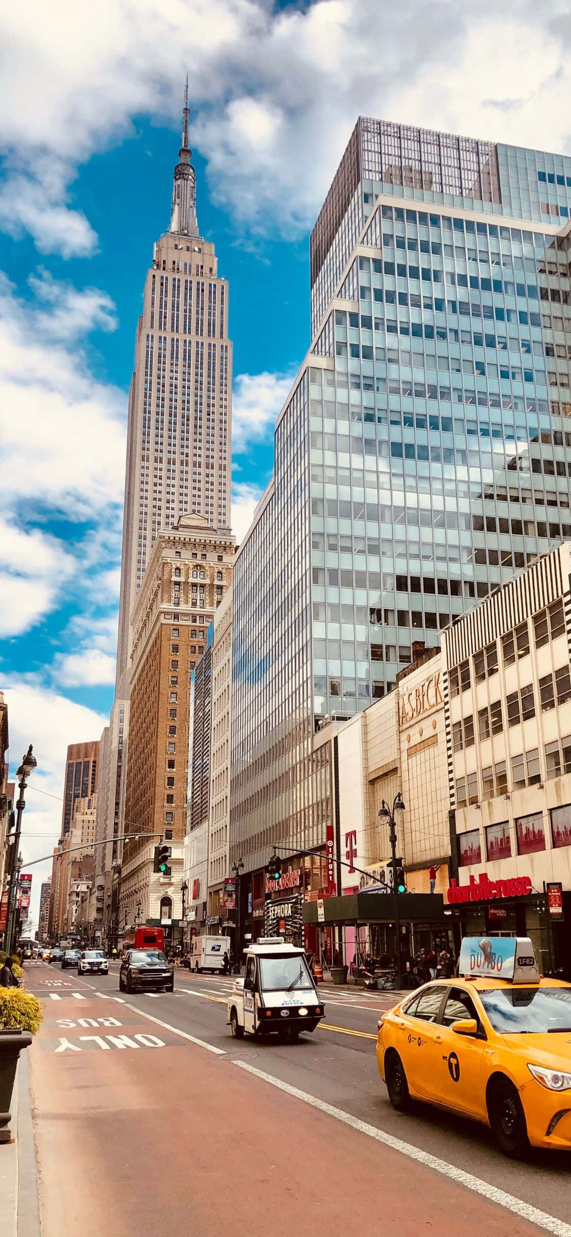 iPhone Xs Ny City 桌布 壁紙 — 充滿活力的紐約市街景，黃色計程車和帝國大廈在明亮的藍天和白雲下高聳入雲。