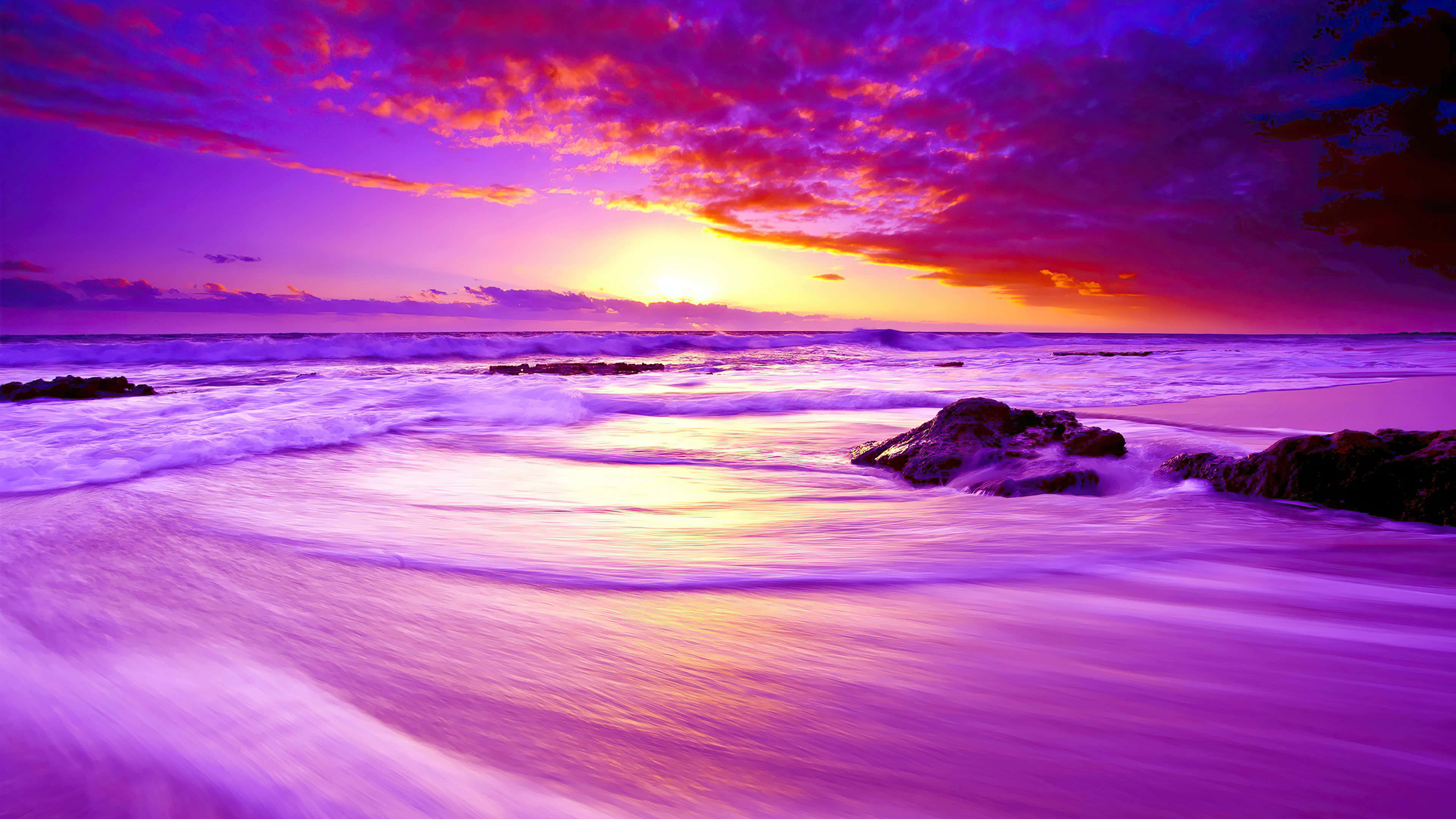 Beach Sunset HD 桌布 壁紙 — 充滿活力的紫色和橙色日落描繪了海浪拍打岩石海岸，上方有戲劇性的雲層。