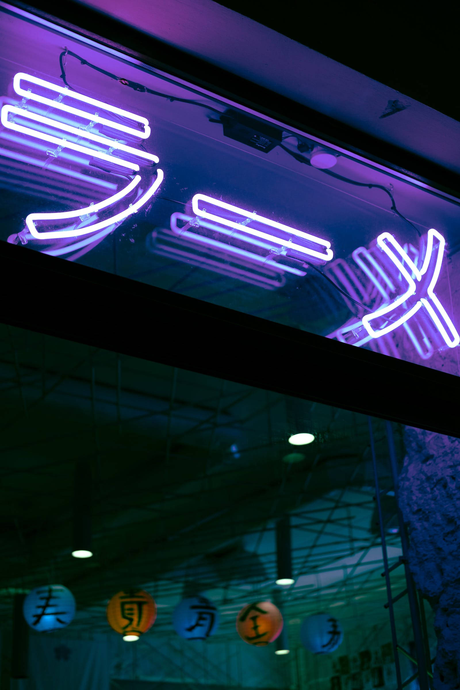 Neon Phone 桌布 壁紙 — 充滿活力的紫色和藍色霓虹燈招牌在黑暗的天花板上發光，天花板上裸露著電線，下方掛著燈籠。