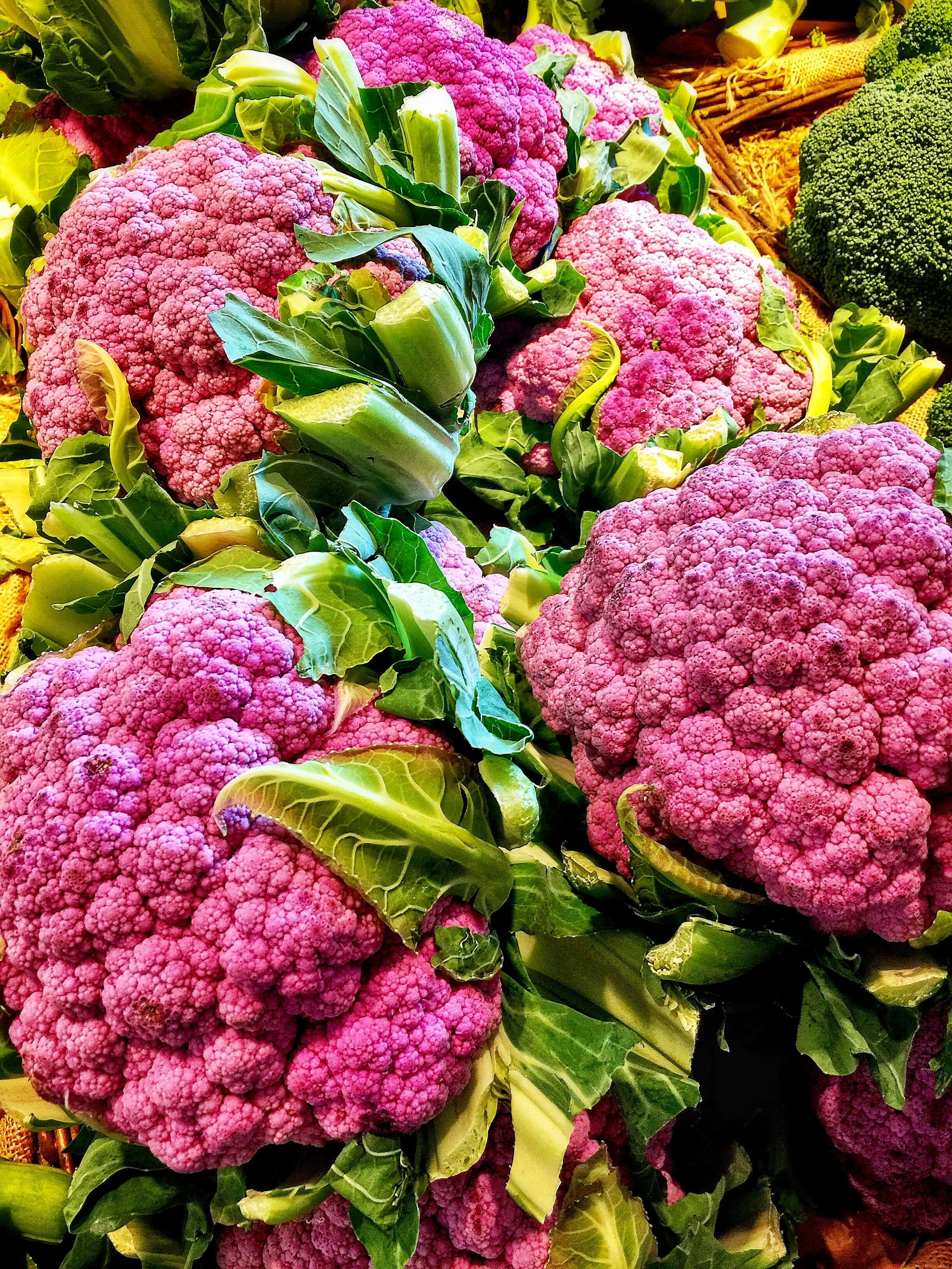 Purple Cauliflower 桌布 壁紙 — 充滿活力的紫色花椰菜頭，搭配茂盛的綠色葉子，堆疊在色彩繽紛的市場展示中。